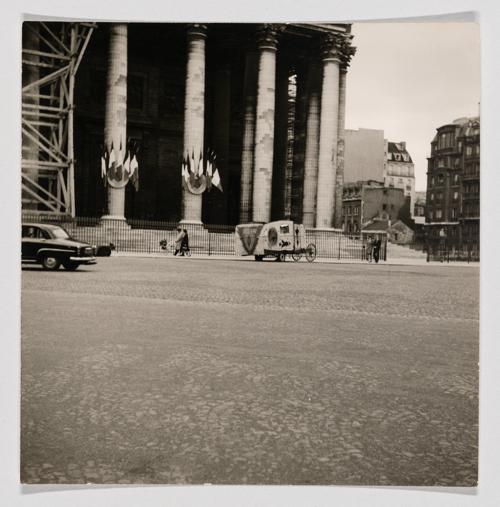Large columned building with flags overlooks a street where a decorated cart and pedestrians pass.