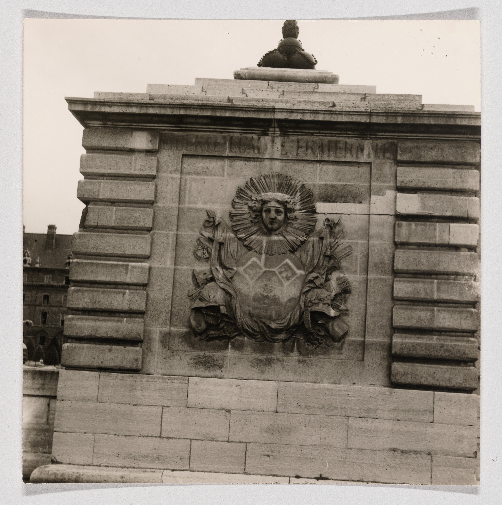 Carved stone relief of a sun-faced figure with radiating rays and draped shield on a masonry wall.