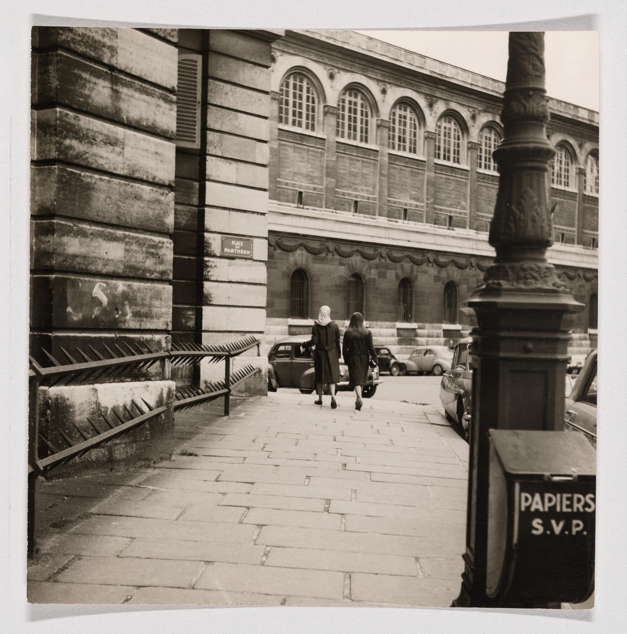 Two women walk down a Paris sidewalk past parked cars and a bin labeled PAPIERS S.V.P.
