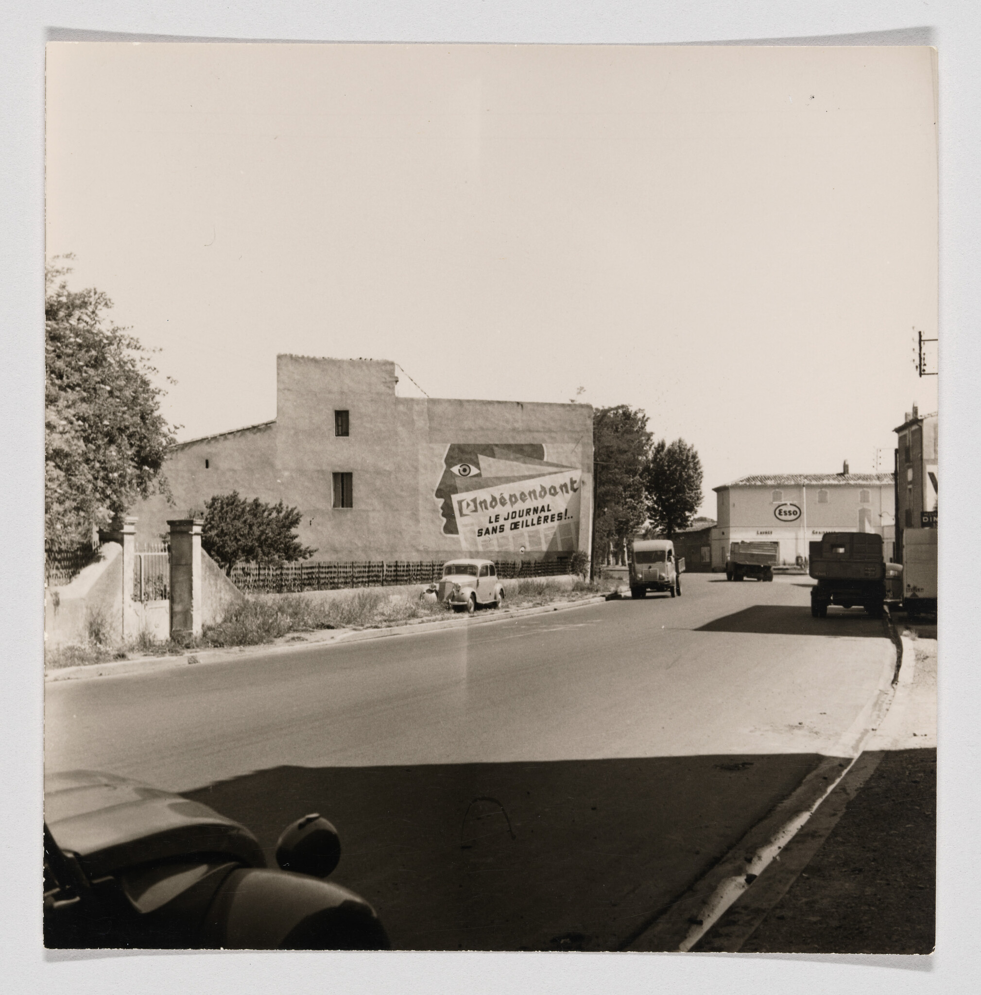 A quiet street with parked vintage cars and a large wall mural advertising "L'indépendant" newspaper.