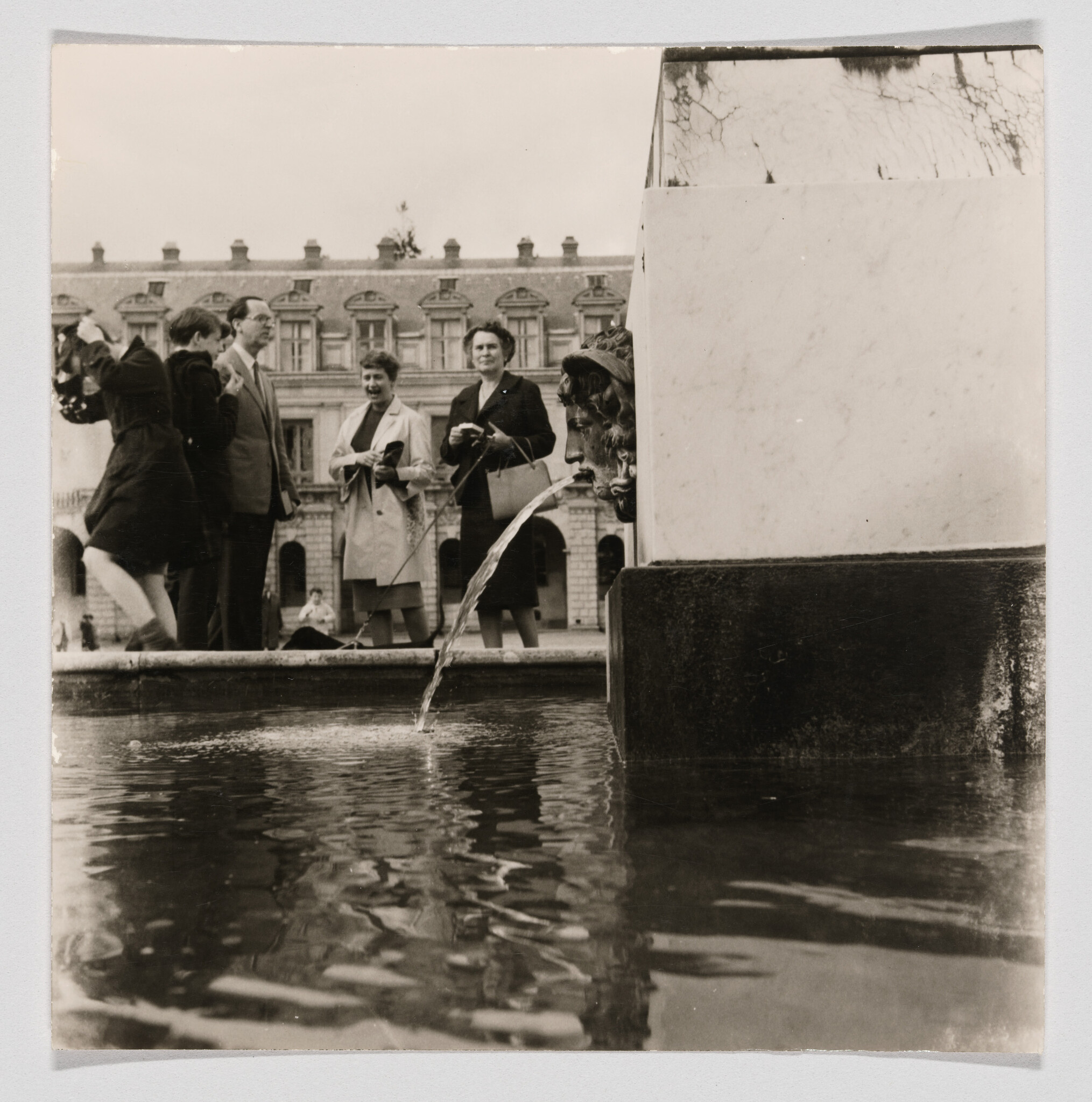 A group of people stand by a plaza fountain as a lion head spout sprays water.