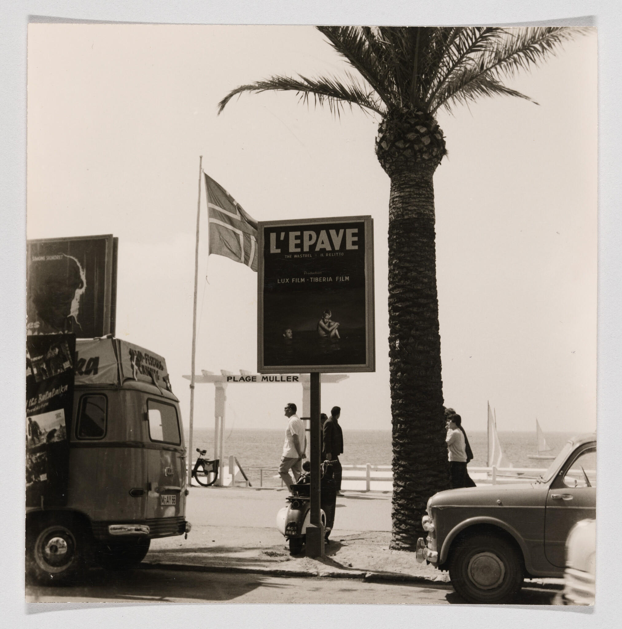 Billboard advertising "L'EPAVE" stands by a palm tree on the seaside promenade with pedestrians.