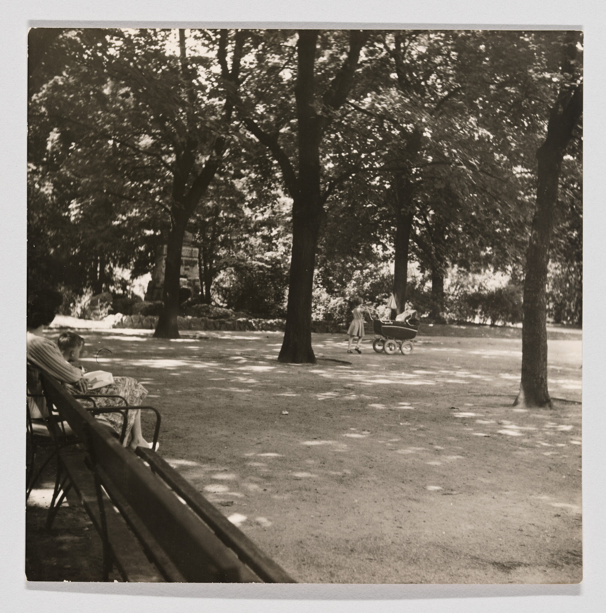 A child pushes a vintage baby carriage across a shaded park while people sit on a bench.