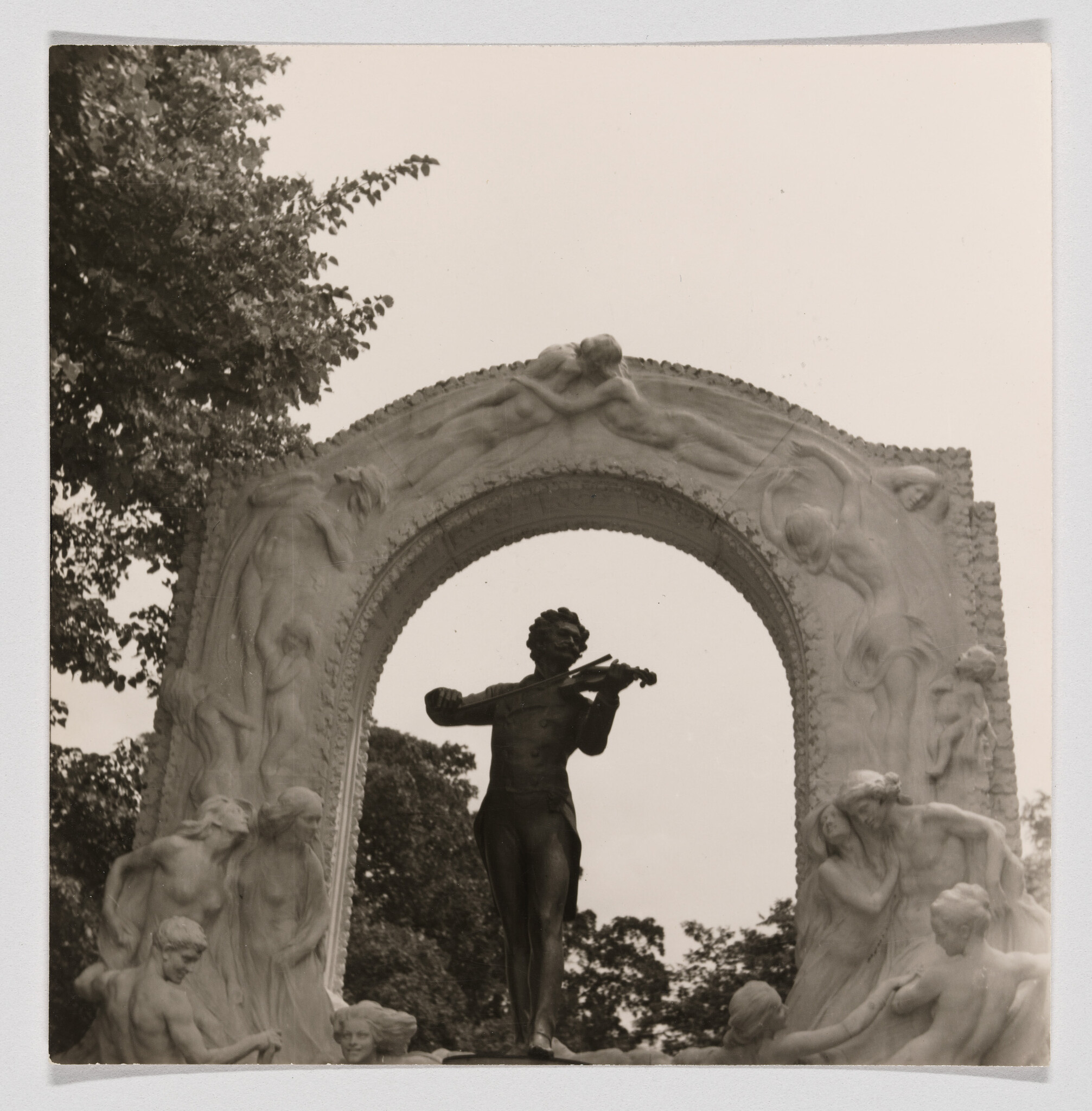 Bronze violinist statue stands under an ornate arch surrounded by relief figures.