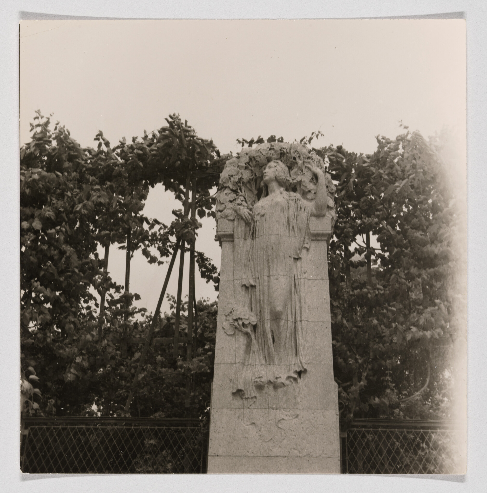 A carved stone female figure in flowing robes stands on a tall pedestal against leafy trellis.