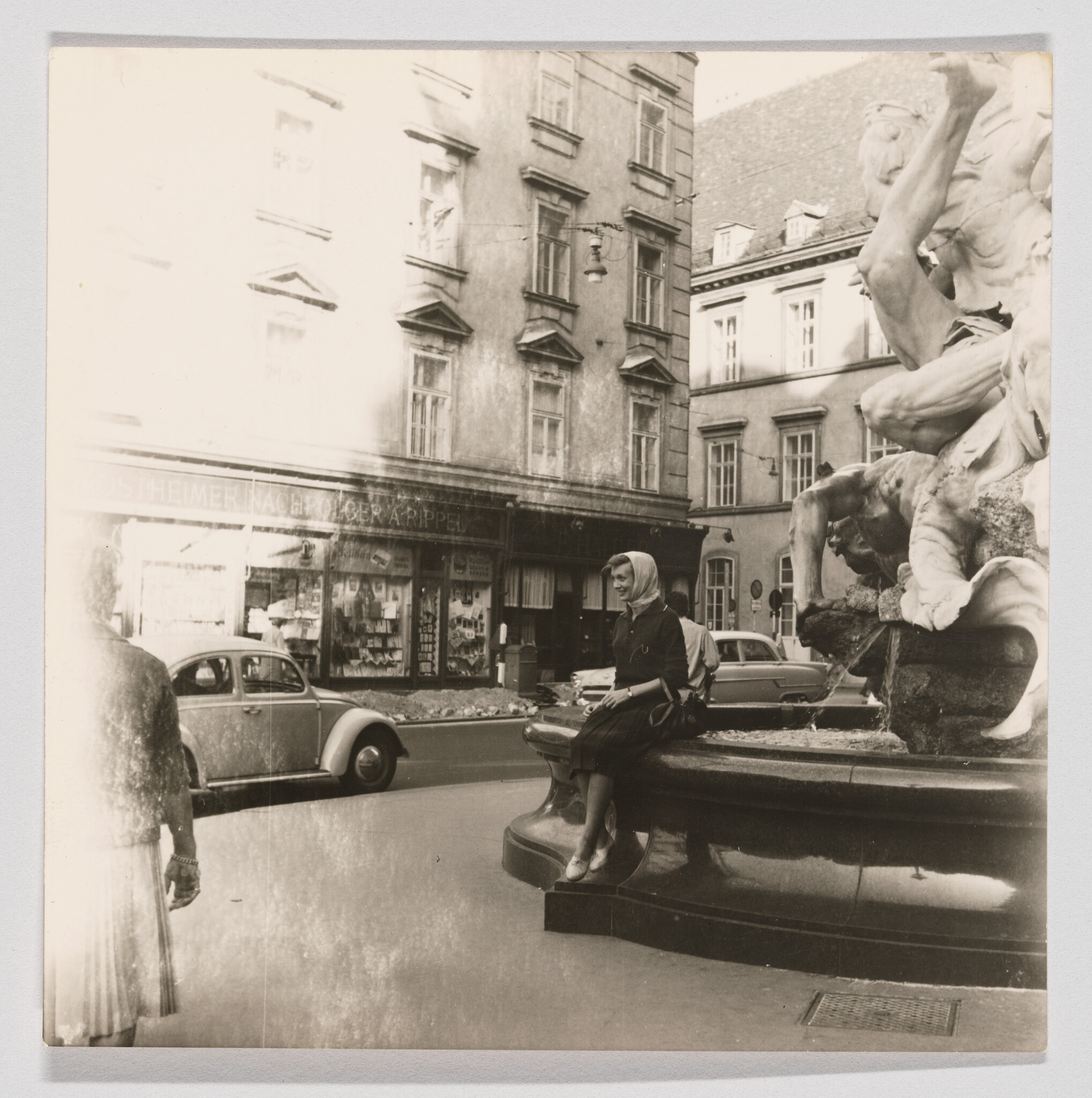 A woman in a headscarf sits on the edge of a fountain in a city square.