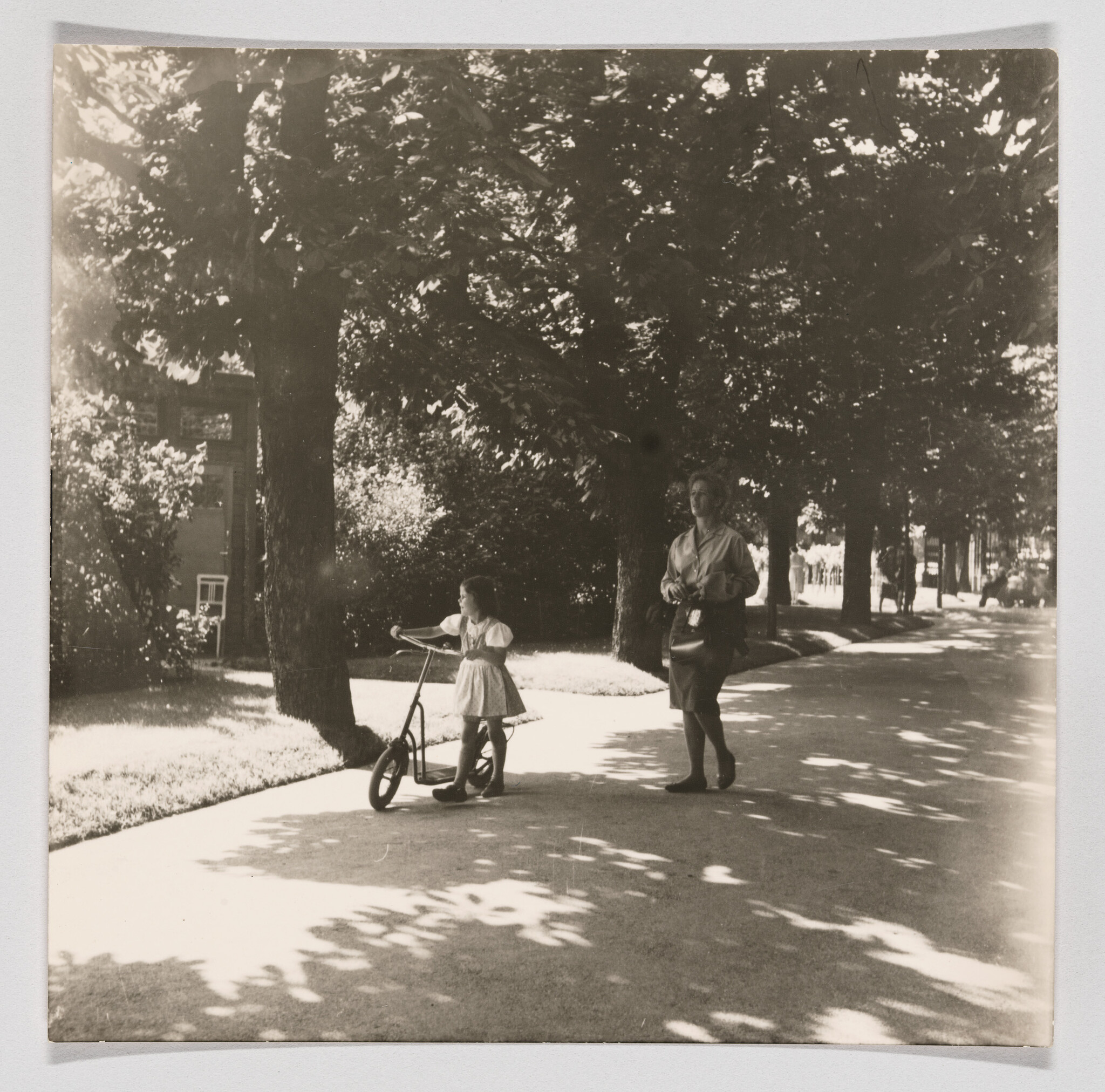A young girl pushes a scooter while a woman walks beside her under shaded trees.