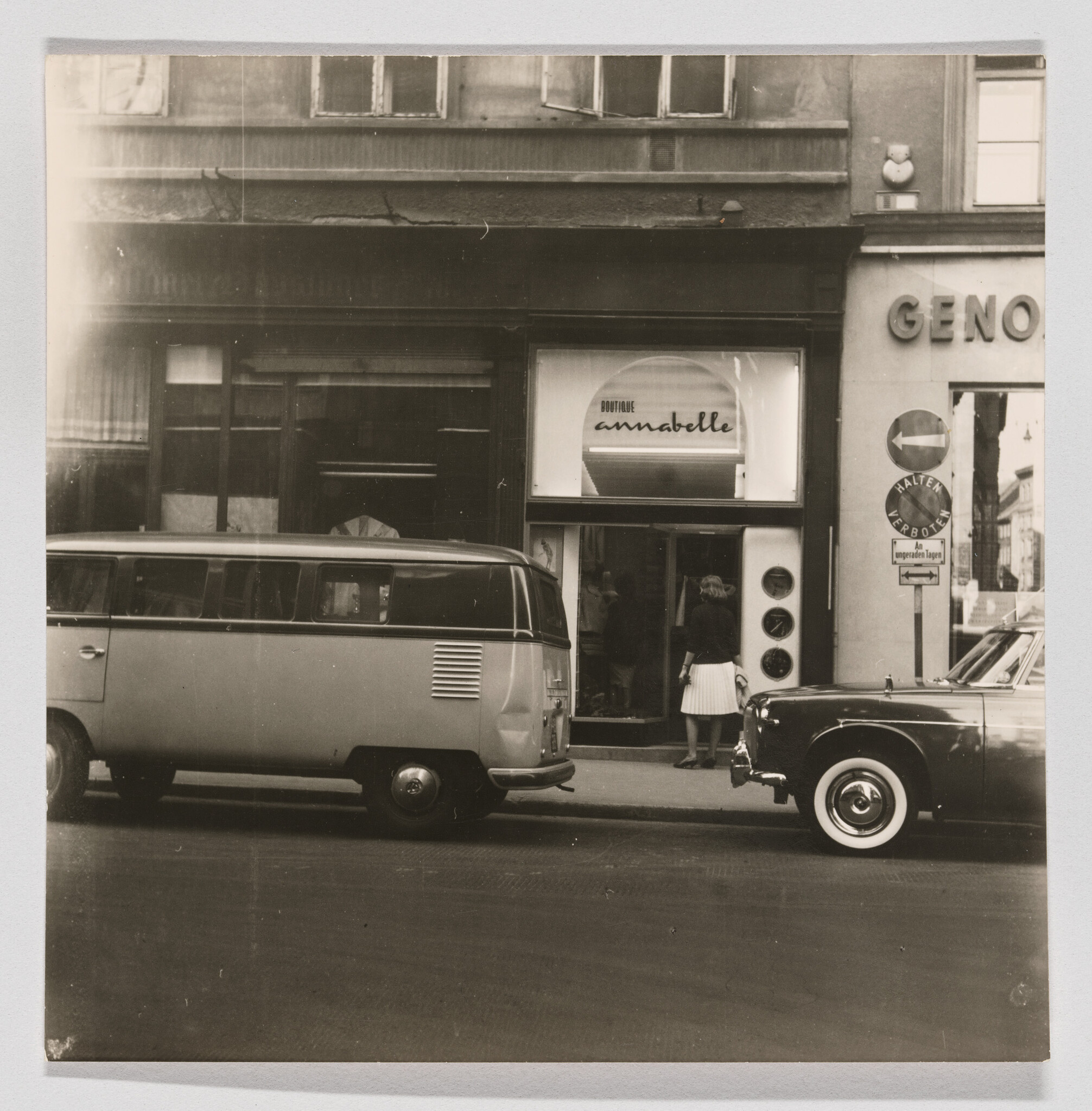 A black and white photograph depicting a vintage street scene with a classic Volkswagen van parked on the left and an old car on the right. In the background, there's a storefront with a sign reading "BOUTIQUE annabelle" and a woman standing at the entrance. The building also has a sign "GENO" on the right side, and there's a traffic sign indicating a right turn only. The image has a nostalgic feel, capturing a moment from a bygone era.