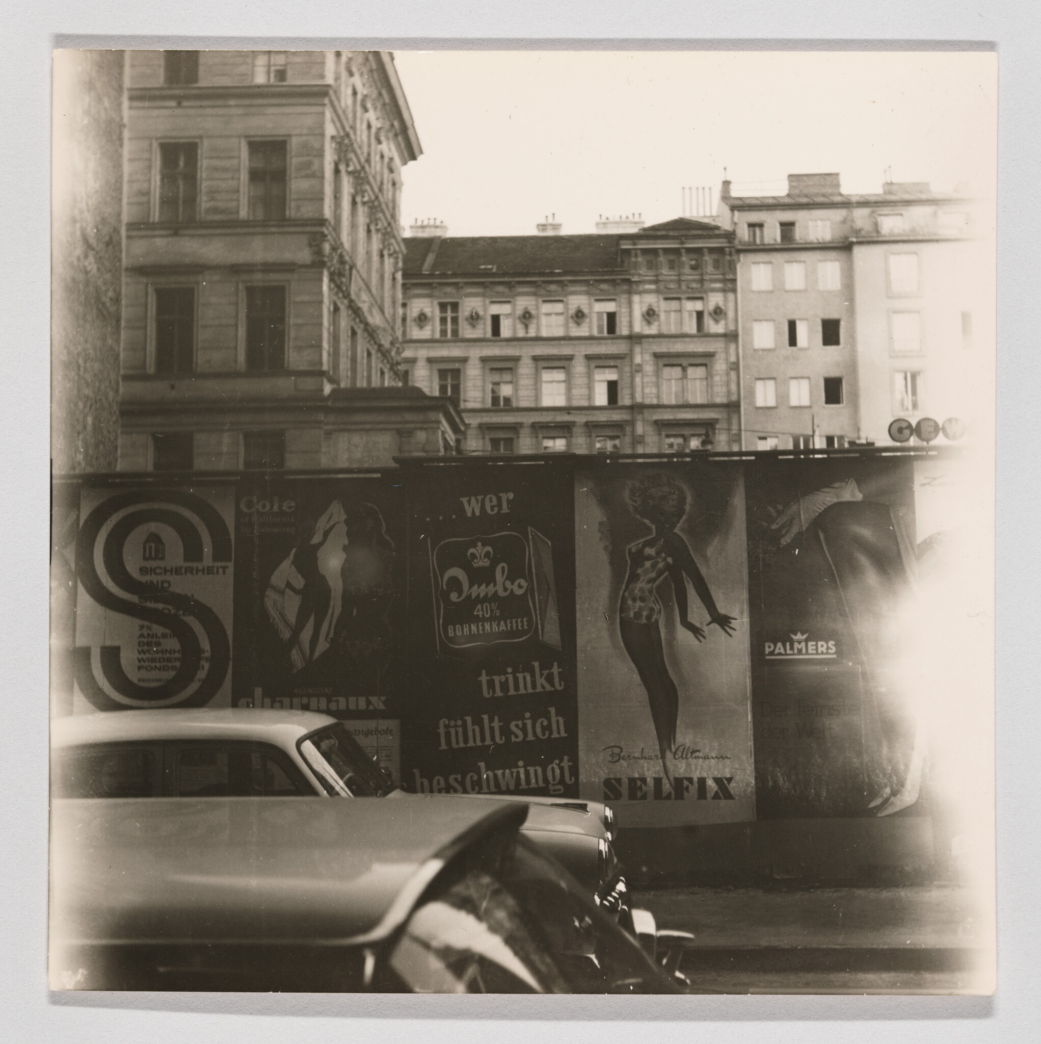 Two vintage cars parked along a city street with advertising posters on a wall behind them.