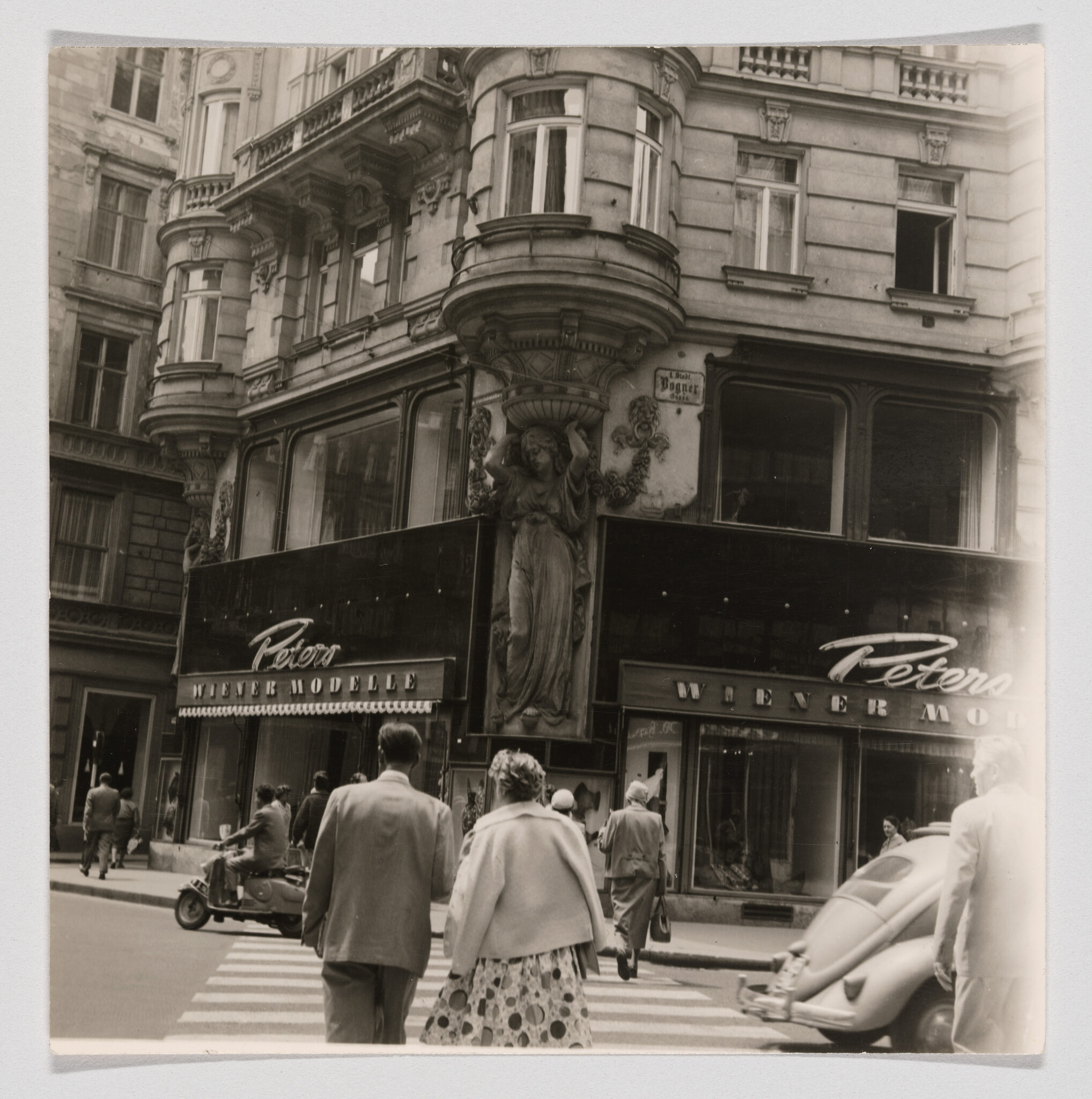 A couple crosses a zebra crossing toward an ornate corner shop facade with a large statue.