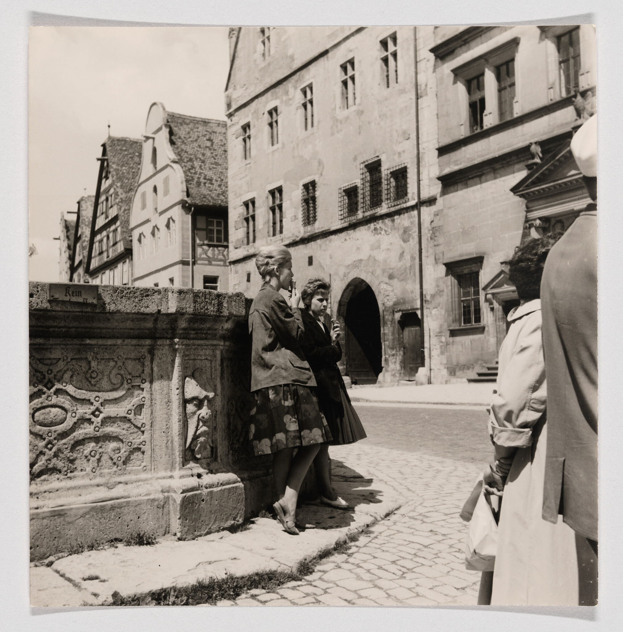 Two young women lean against a carved stone fountain eating ice cream in a quiet cobbled town square.