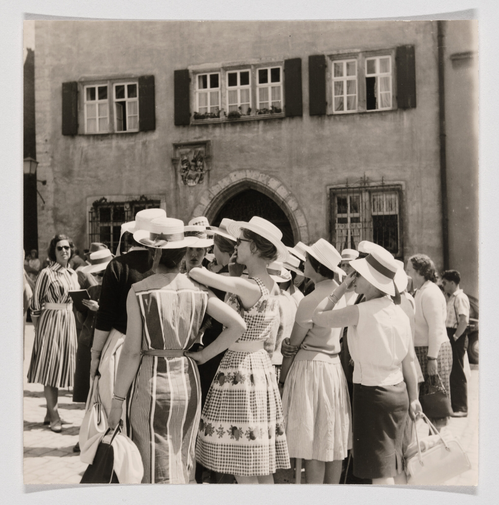 A black and white photograph depicting a group of women wearing summer dresses and wide-brimmed hats gathered in a sunny square in front of an old building with shuttered windows and an arched doorway. Some of the women are engaged in conversation, while others look off into the distance. The attire and quality of the image suggest a mid-20th-century setting.