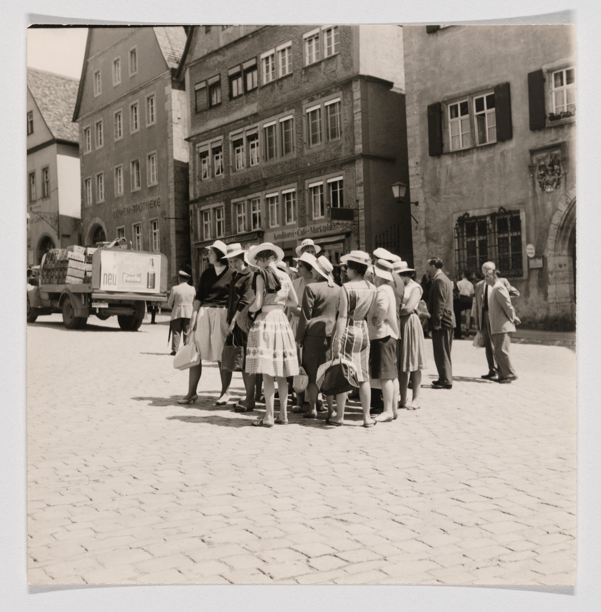 A group of women in hats stand together on a cobblestone town square in front of old buildings.
