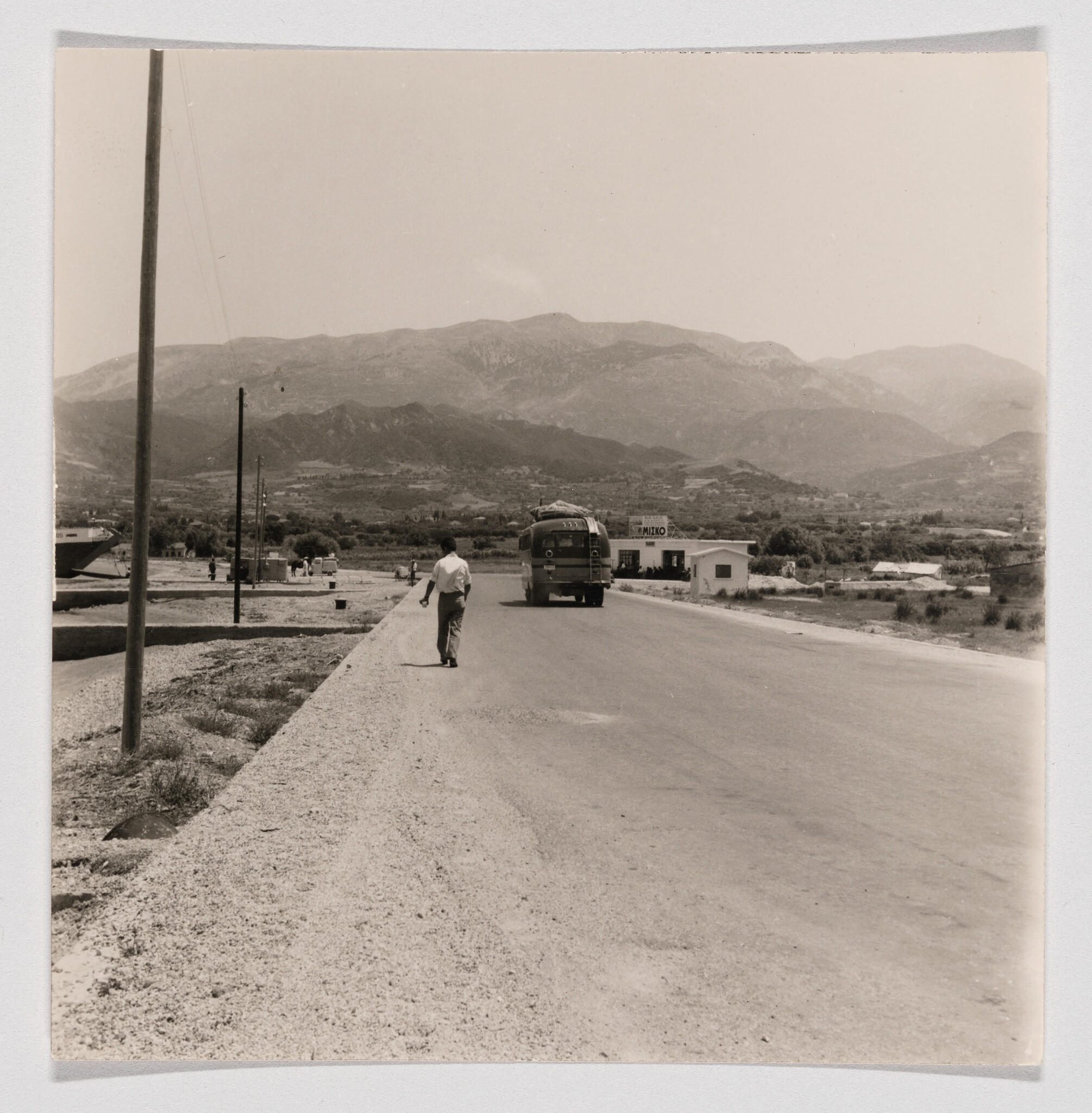 A man walks along a rural road toward a bus and distant mountains under a clear sky.