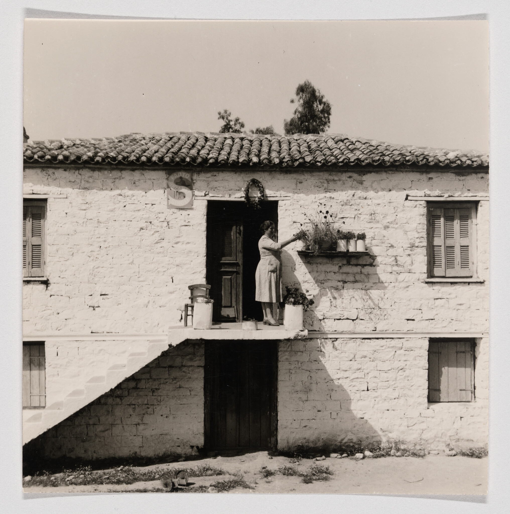 A woman stands on an upper balcony tending potted plants outside a white stone house.