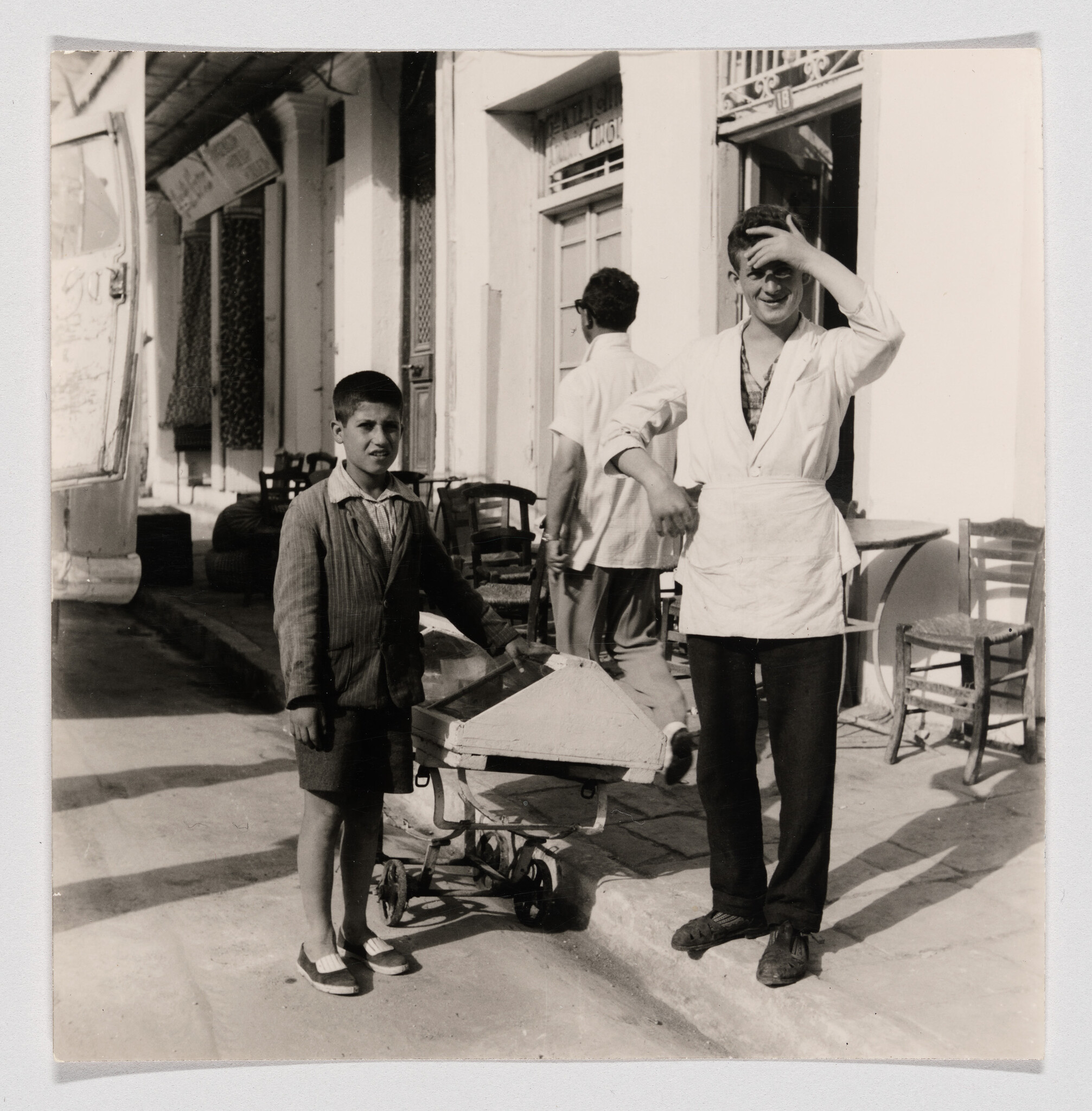 Young boy pushes a small wooden cart while a man in an apron shields his eyes from the sun.