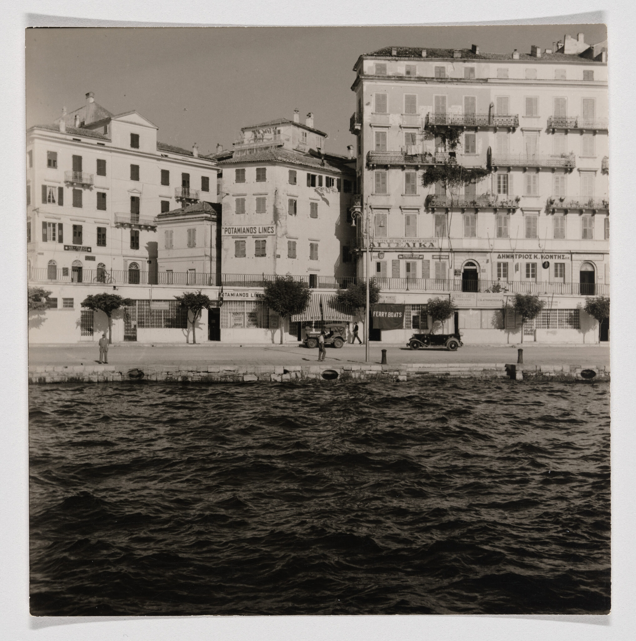 A vintage black and white photograph showing a waterfront scene with a row of multi-story buildings. The buildings have balconies and signage, including one that reads "POTAMIANOS LINES." In front of the buildings, there is a promenade with a few parked cars and a couple of pedestrians. The foreground features choppy water, suggesting the photo was taken from a boat or across the water.