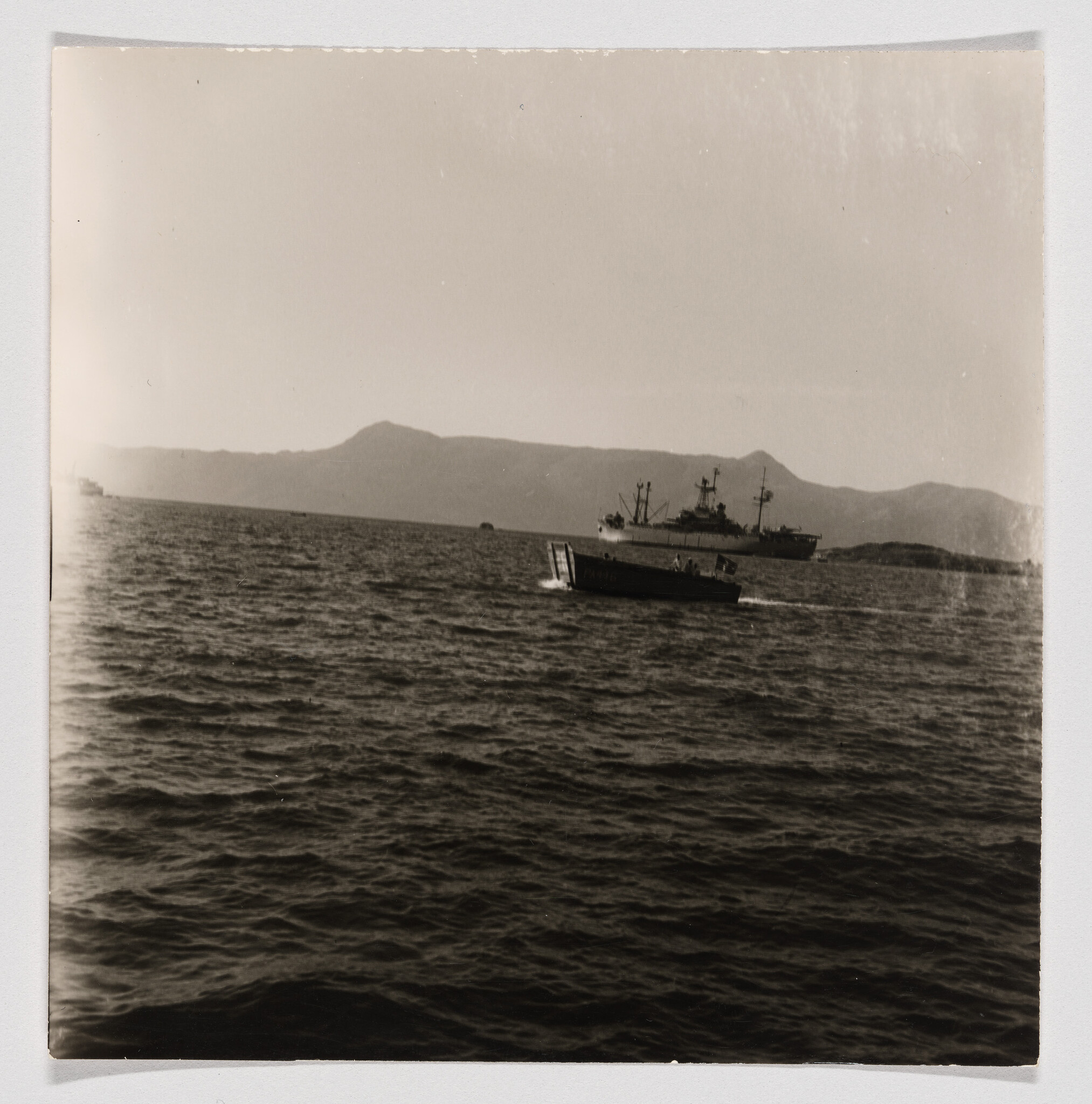 A small motorboat speeds across choppy water toward a large anchored ship with distant mountains.