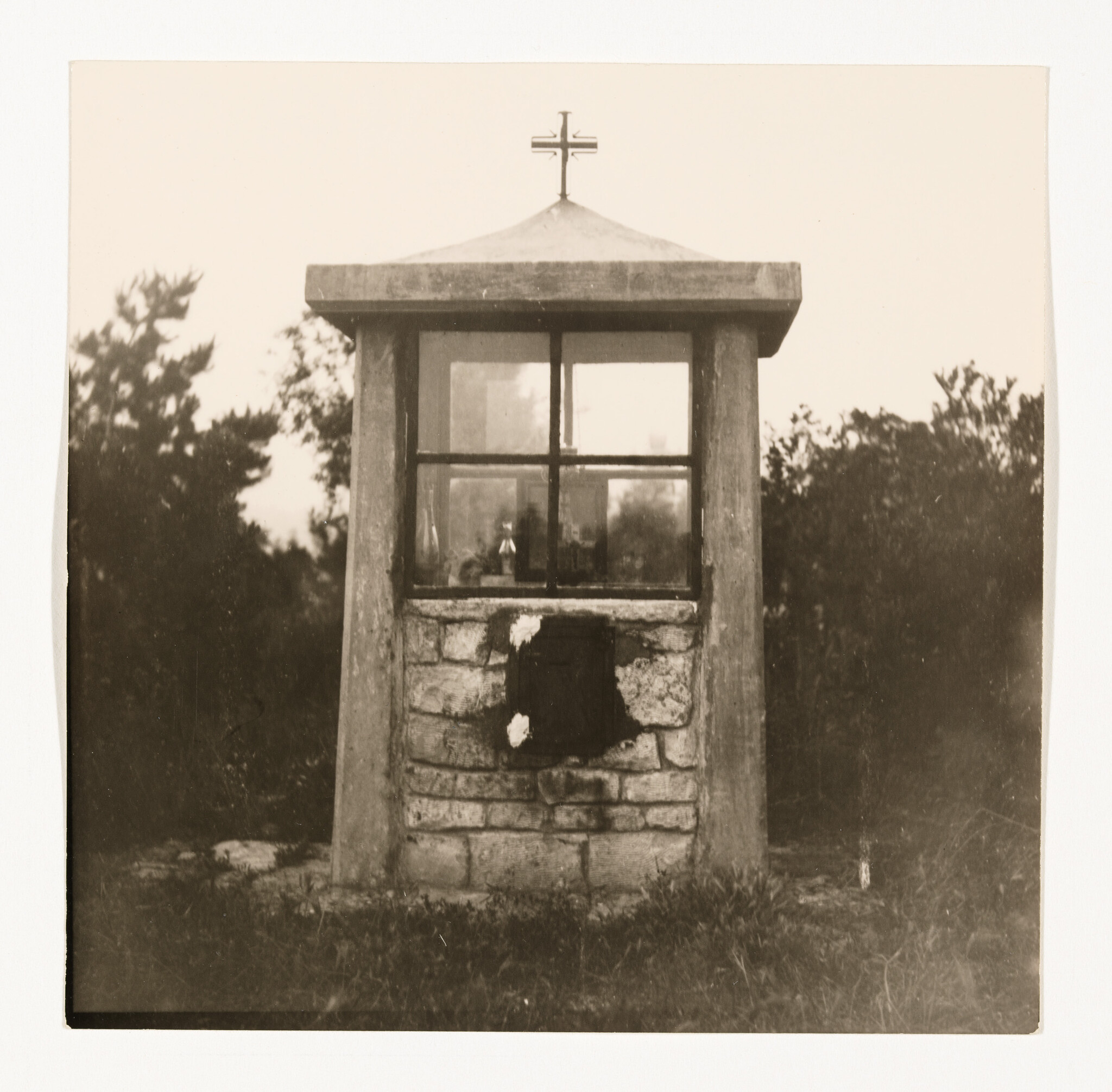 Small stone roadside shrine with glass window and cross on top standing in grass.