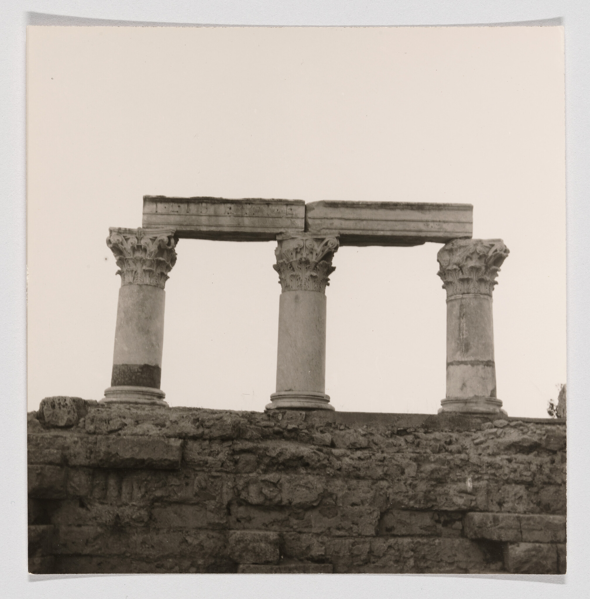 Three ancient Corinthian columns with decorative capitals stand atop a rough stone ruin wall.