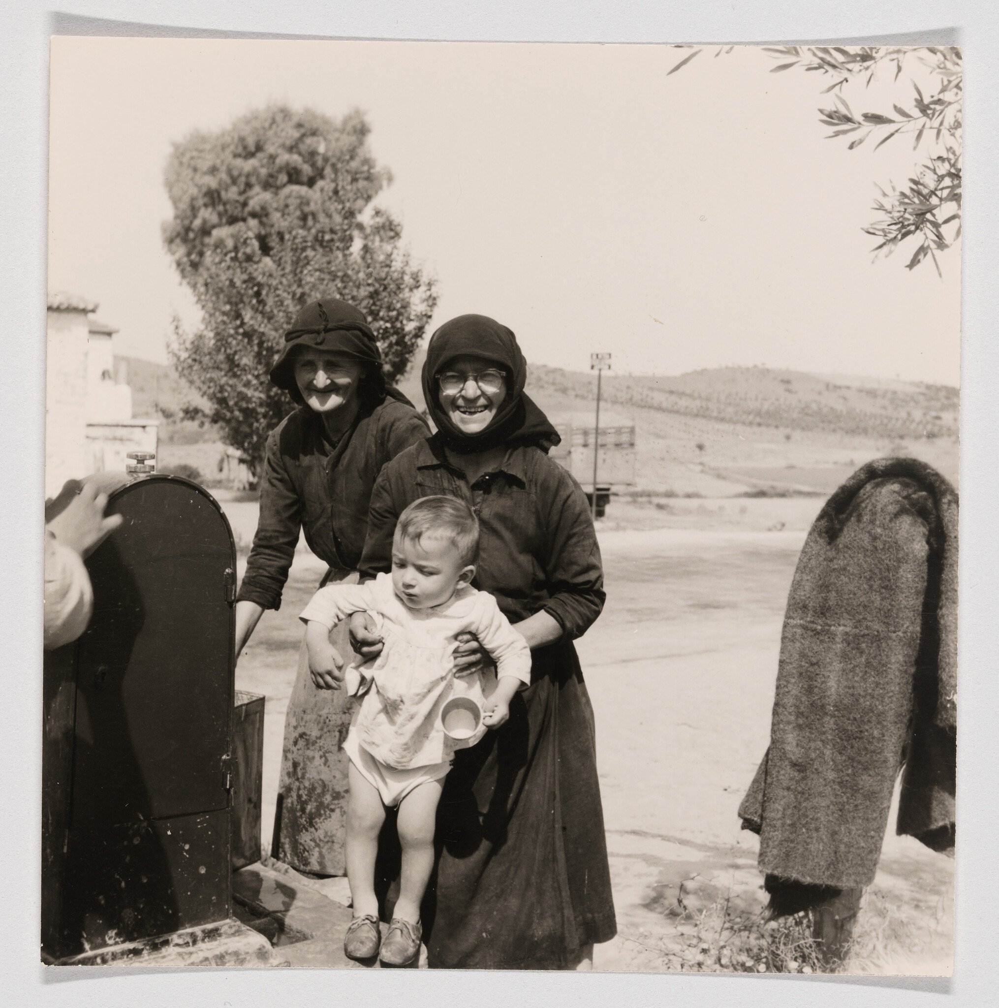Two elderly women smiling as they hold a young child outdoors near a roadside.