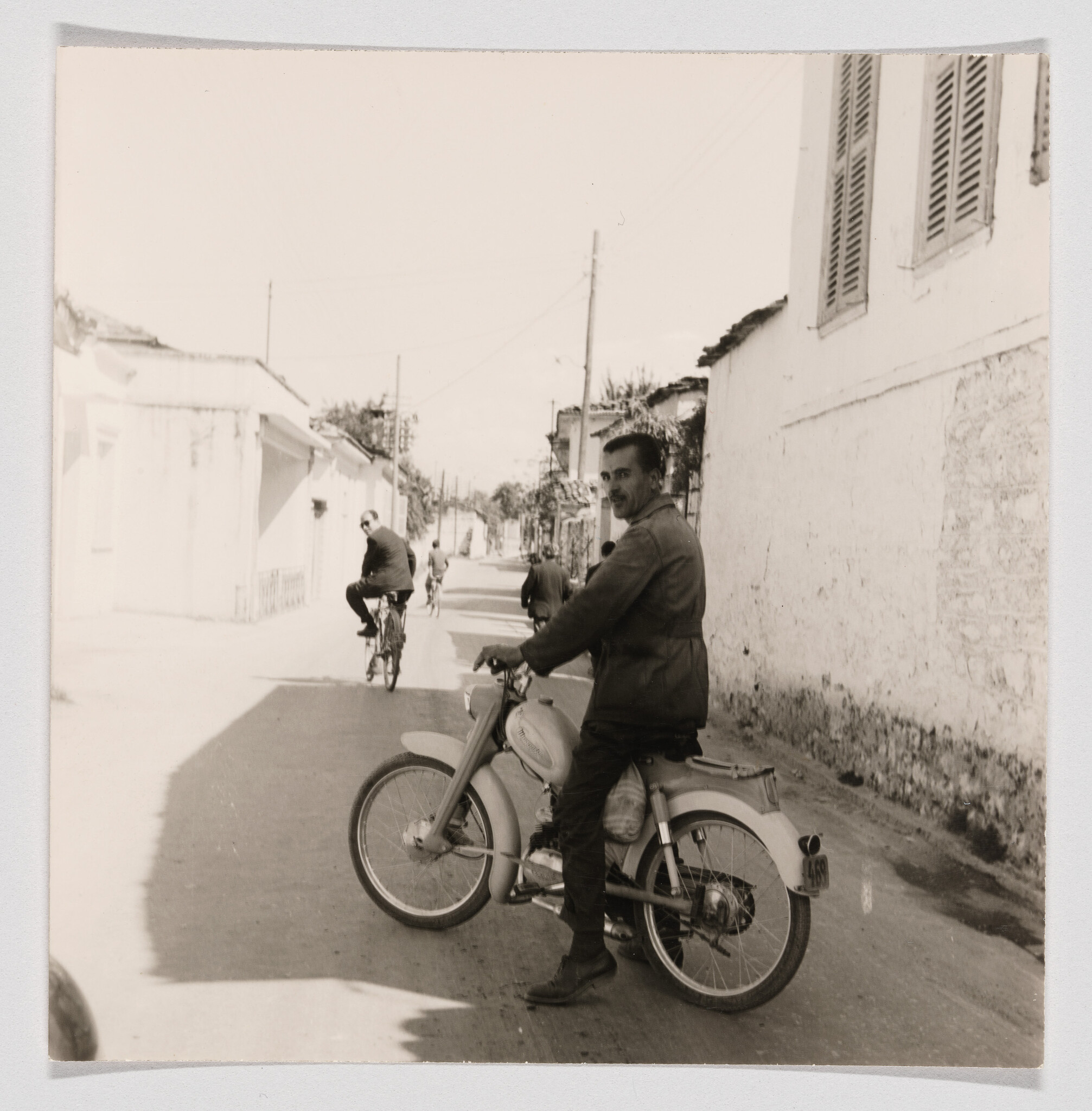 A black and white photograph capturing a moment on a quiet street where a man is seated on a stationary motorcycle, looking over his shoulder at the camera. In the background, another individual is riding a bicycle away from the viewer. The setting appears to be in a residential area with houses lining the street and utility poles visible in the distance. The image has a vintage feel, suggesting it may have been taken several decades ago.