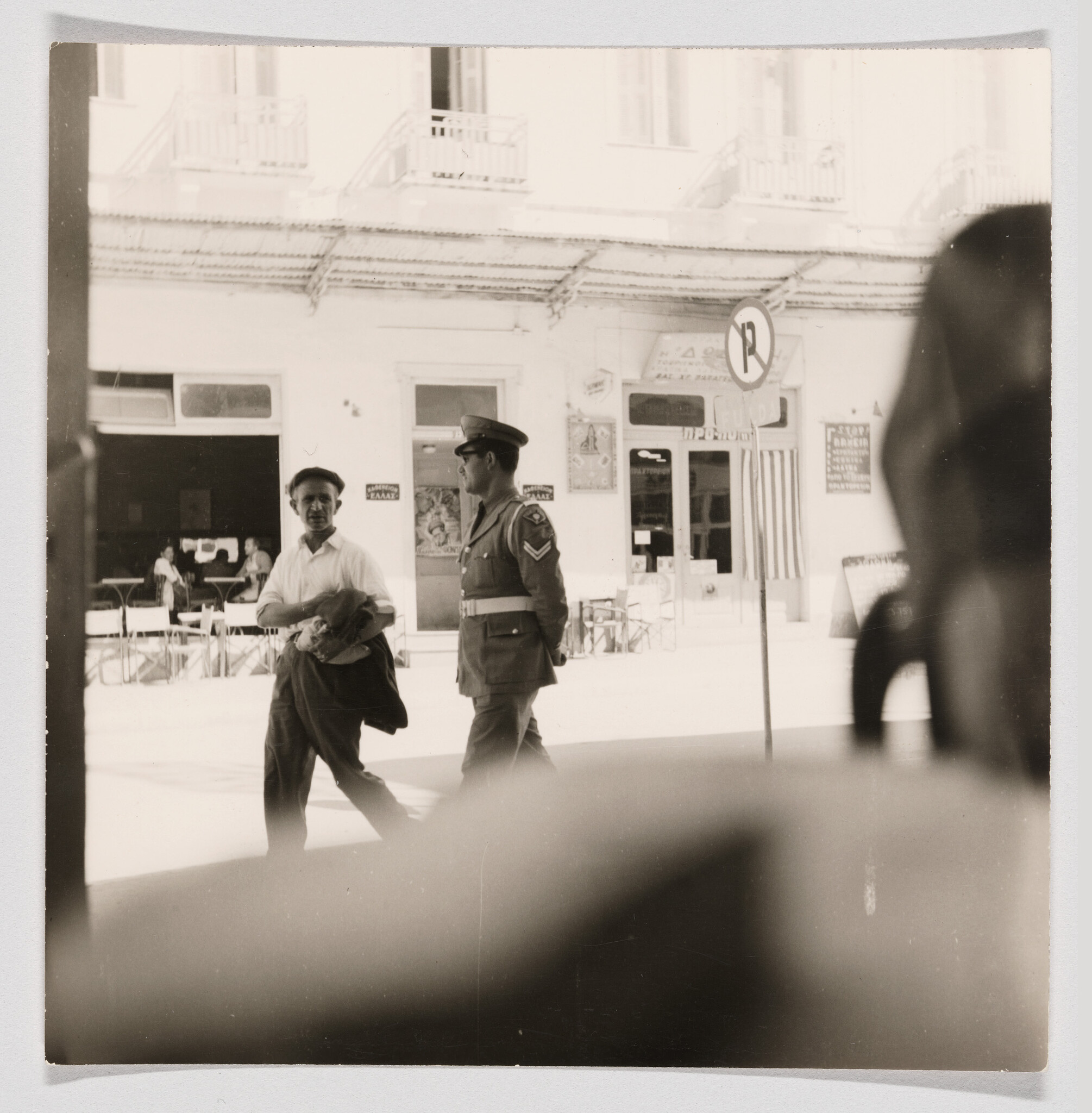 Uniformed police officer walks past a man carrying a jacket on a sunny street.