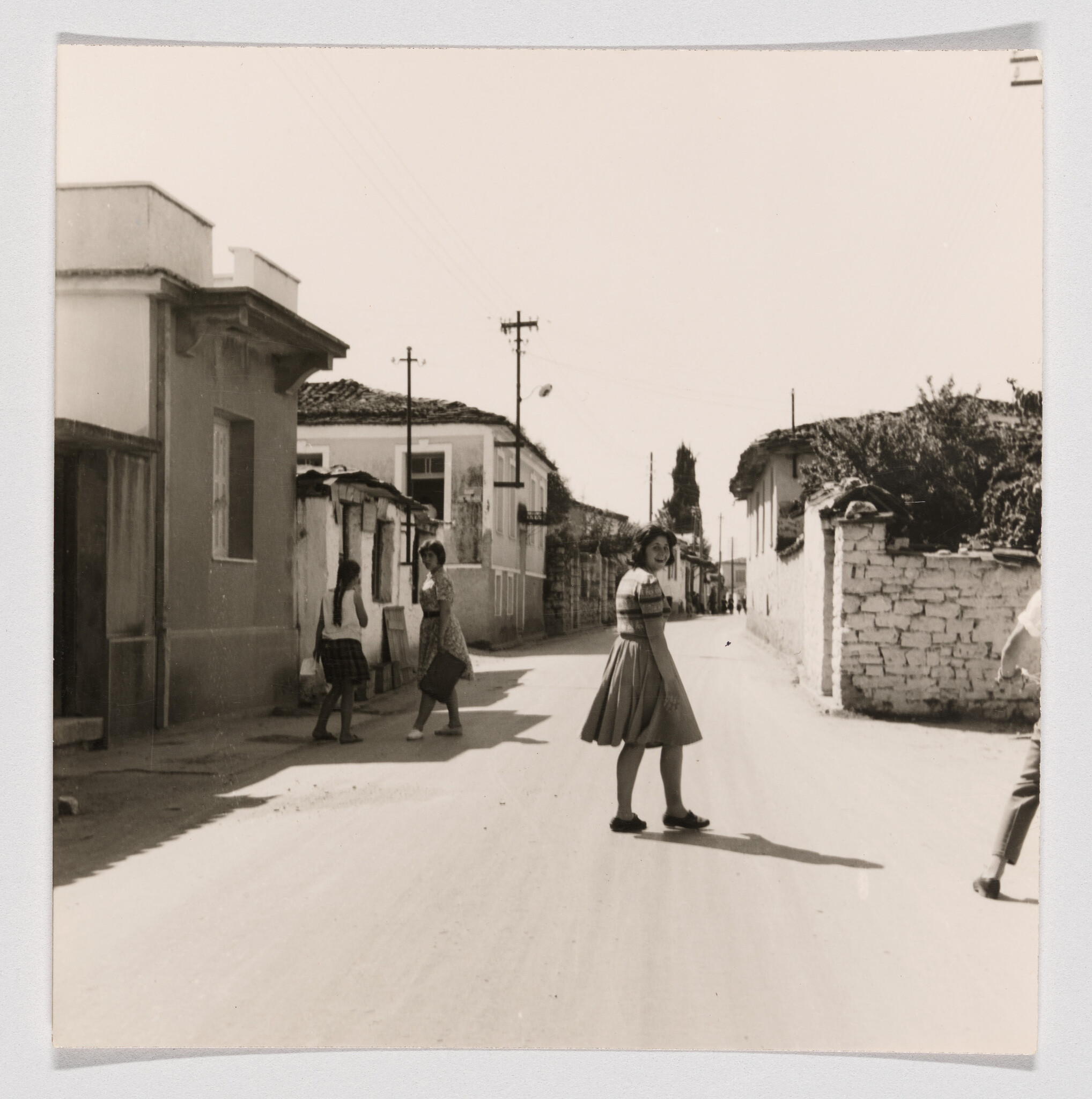 A teenage girl crosses a sunlit village street looking back while other girls stand nearby.