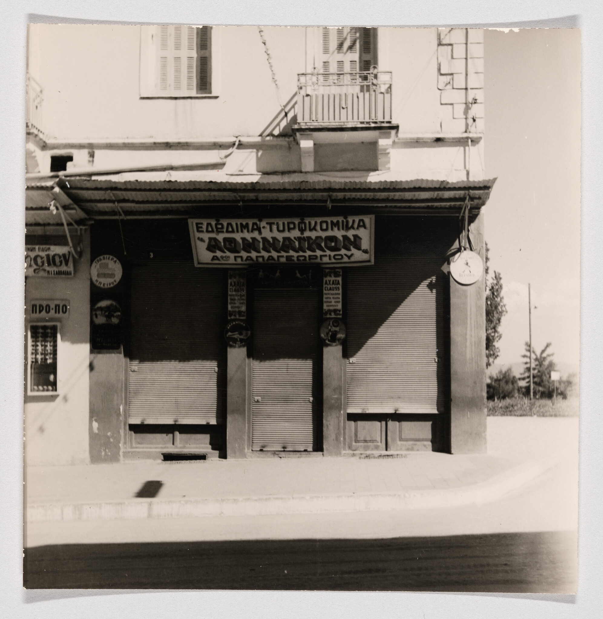 Closed storefront with Greek sign and metal roll-up shutters under a building balcony.