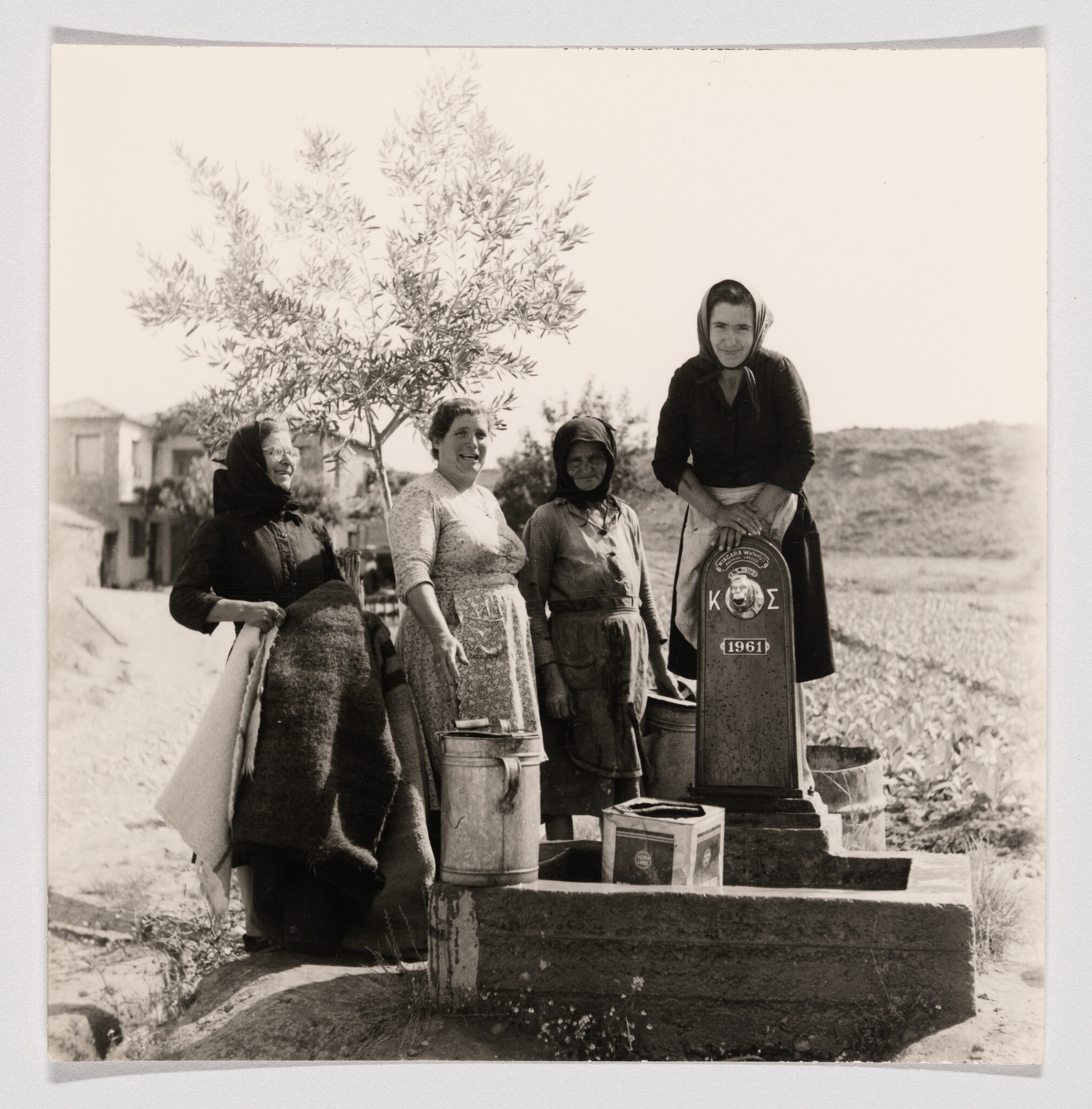 Four women gather water at a public pump, carrying buckets and blankets, 1961.
