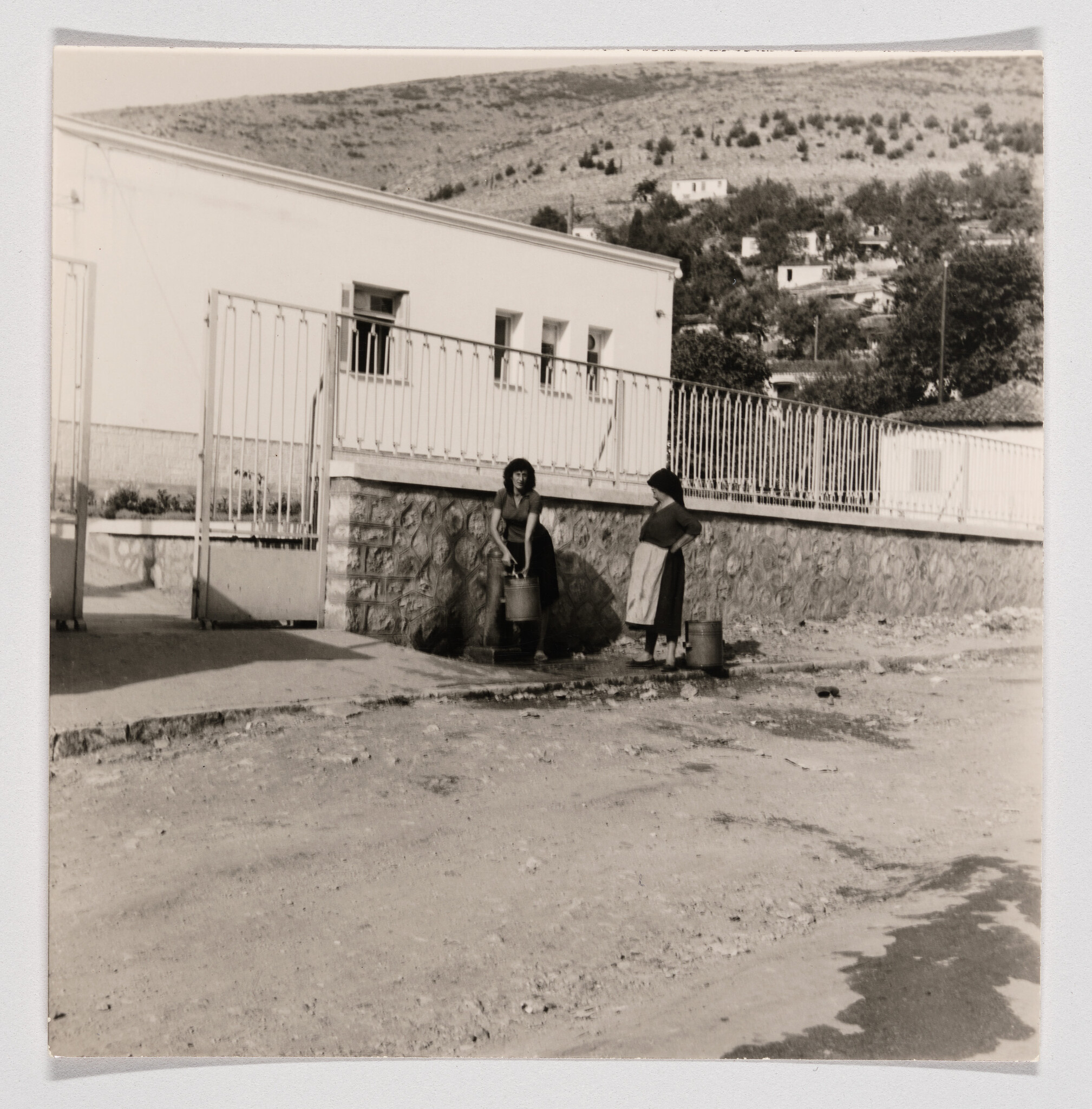 Two women fill metal buckets at a public water tap beside a stone wall and building.