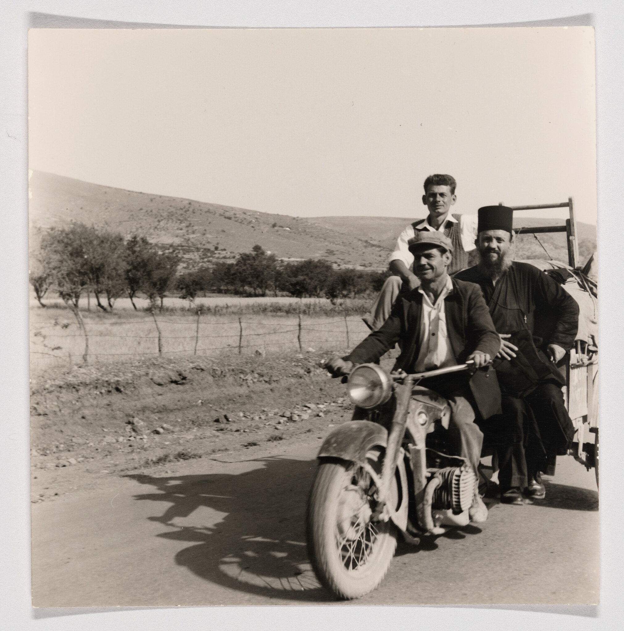 Three men ride a motorcycle and small trailer along a rural road, one wearing clerical robes.