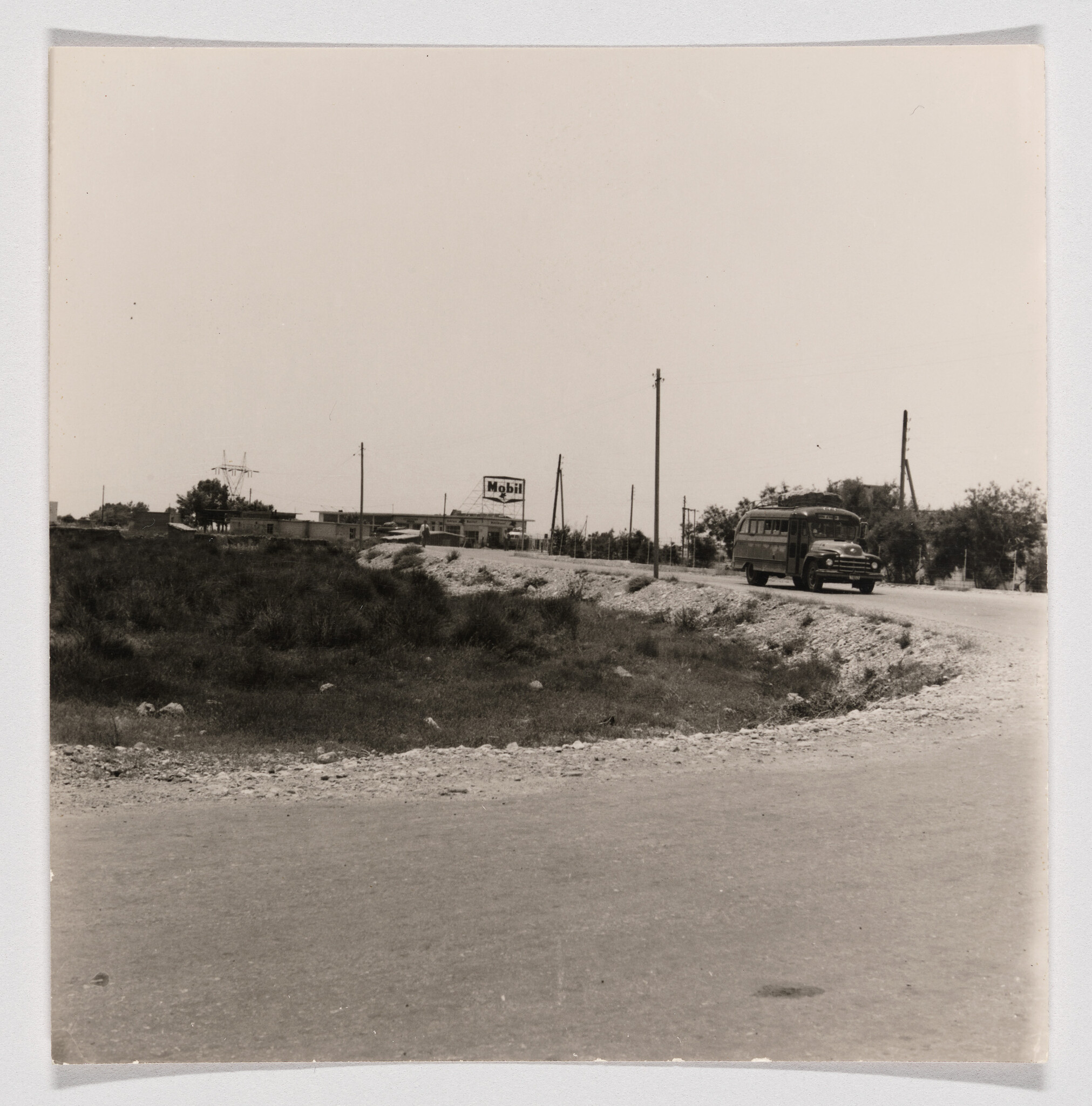 A vintage bus drives along a rural road past a Mobil sign and utility poles.