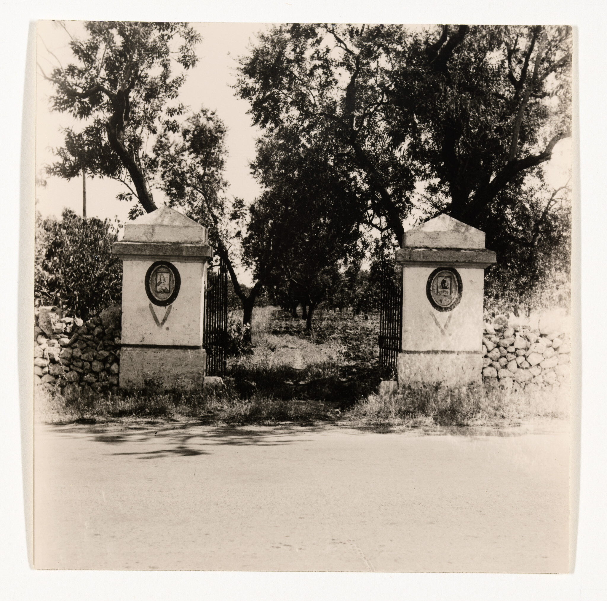 Two stone pillars with emblems flank an open iron gate leading to a tree-lined dirt path.