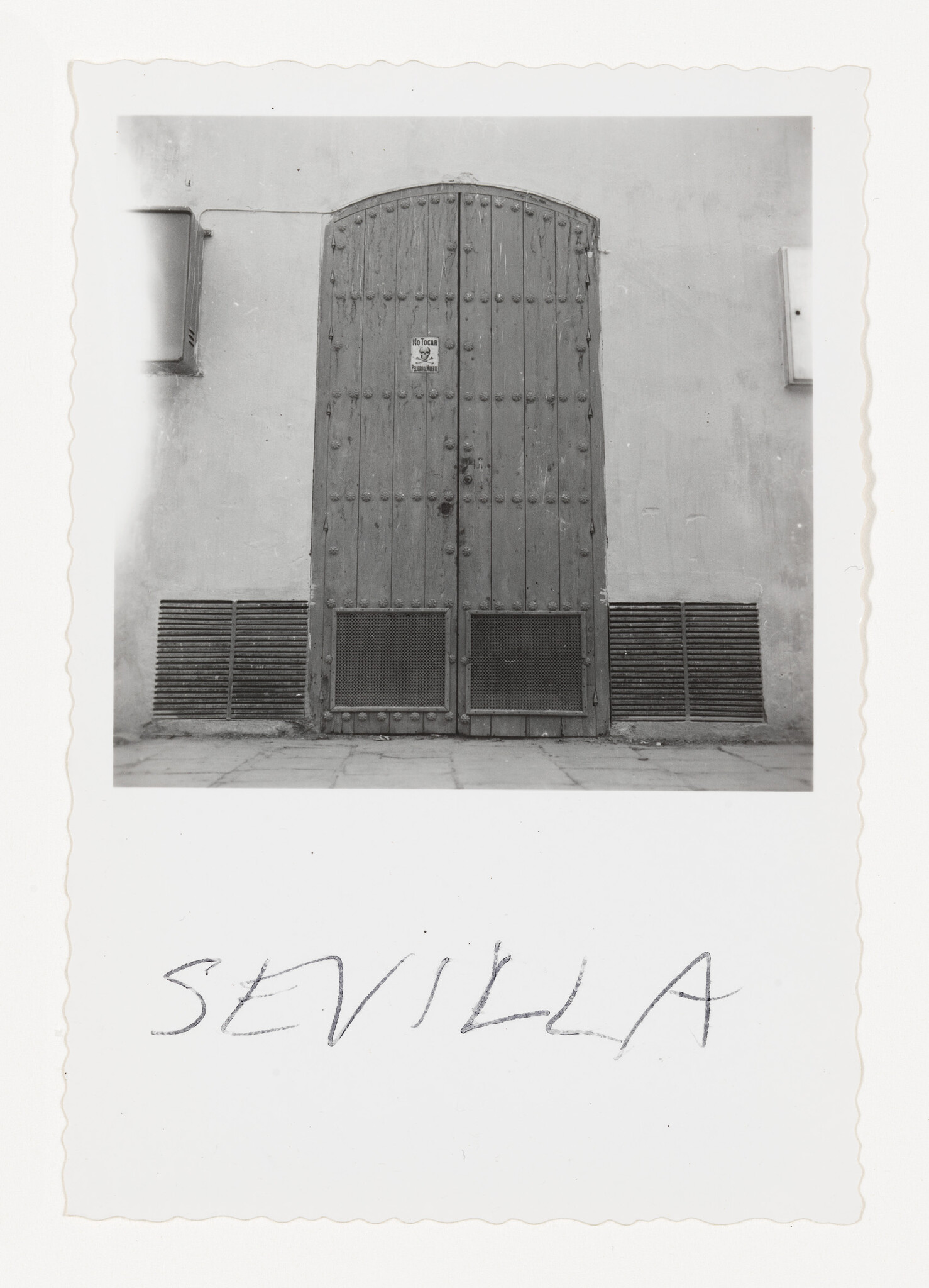 A tall weathered wooden arched door in a plain wall with handwritten "Sevilla" below.