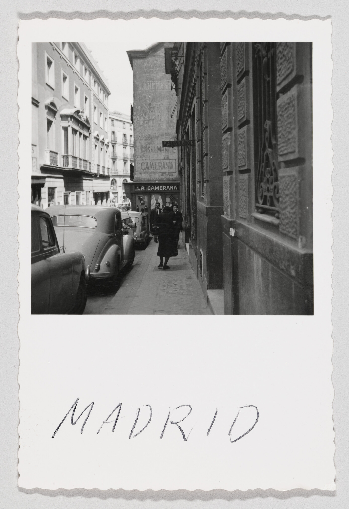 A woman walks on a narrow Madrid sidewalk past parked cars and a "La Camerana" sign.