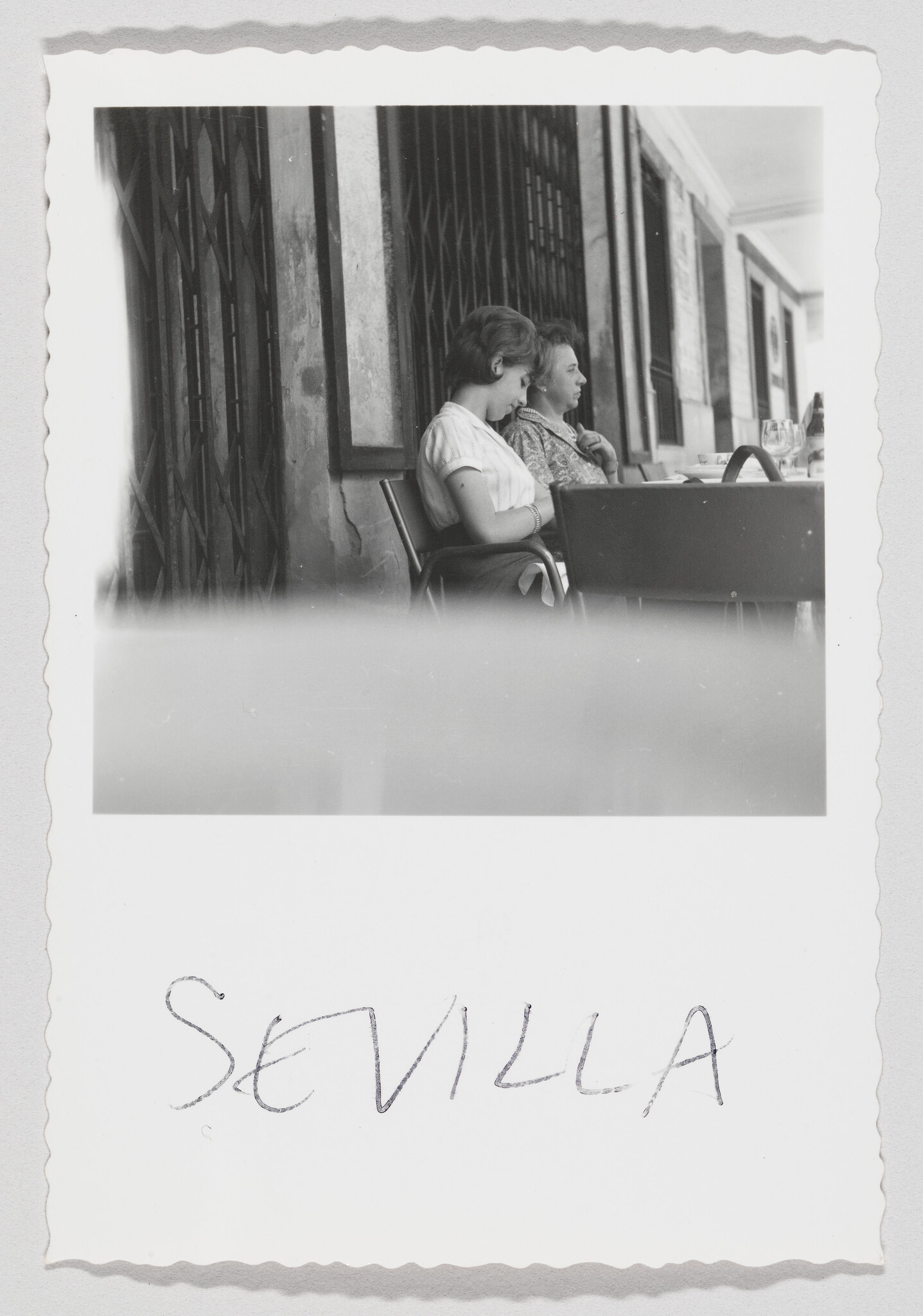 Two women sit at a cafe table on a covered sidewalk, with "Sevilla" handwritten below.