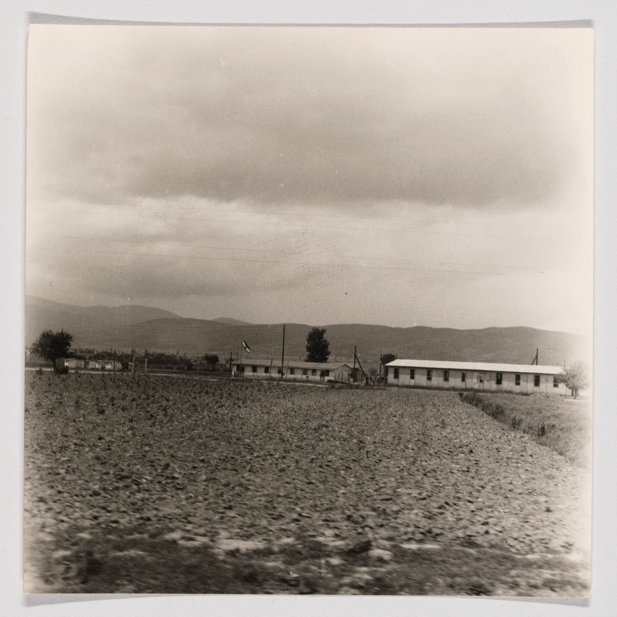A plowed field in front of single-story buildings with mountains and a cloudy sky.