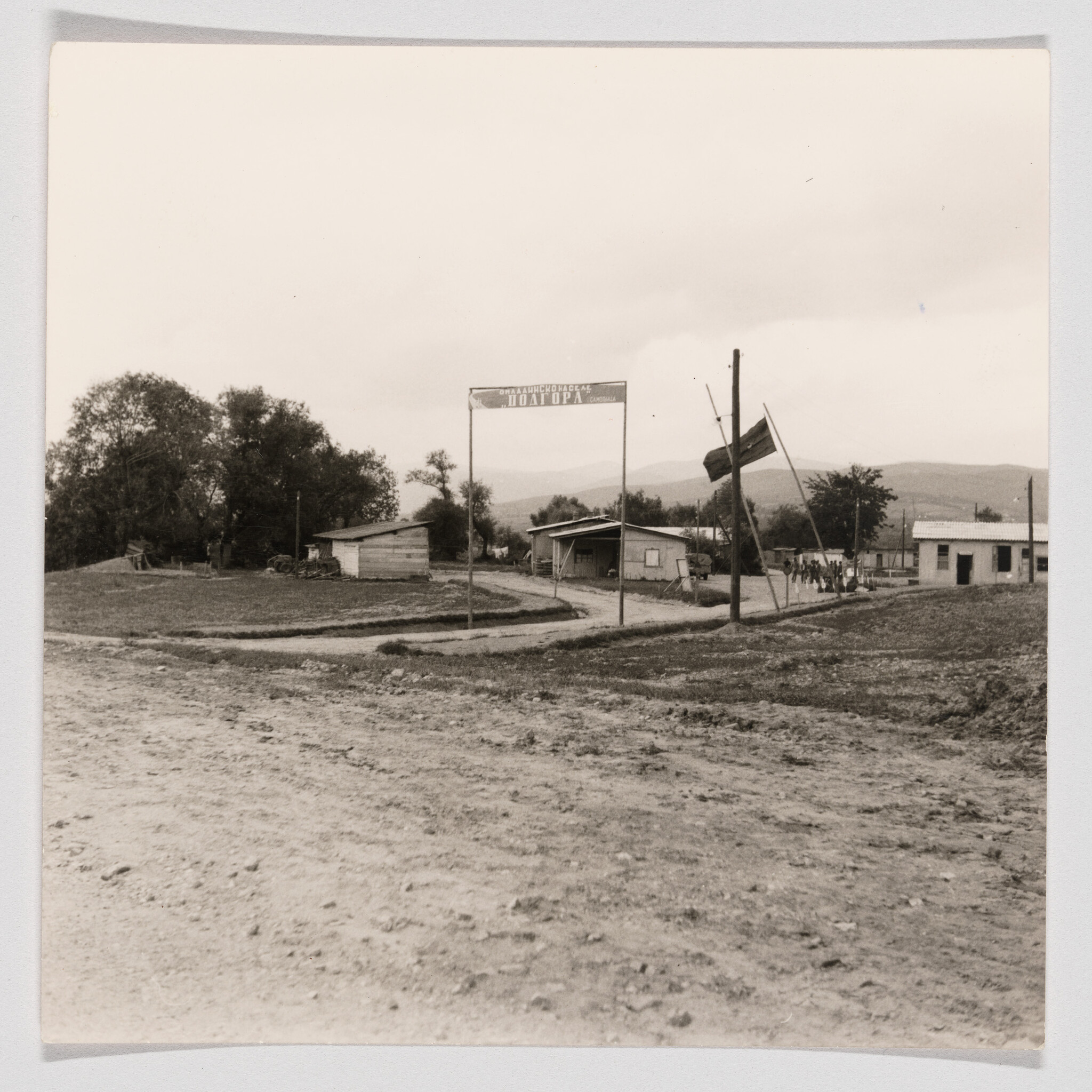 Dirt road leading to a small rural camp entrance with a sign, flagpole, and simple buildings.