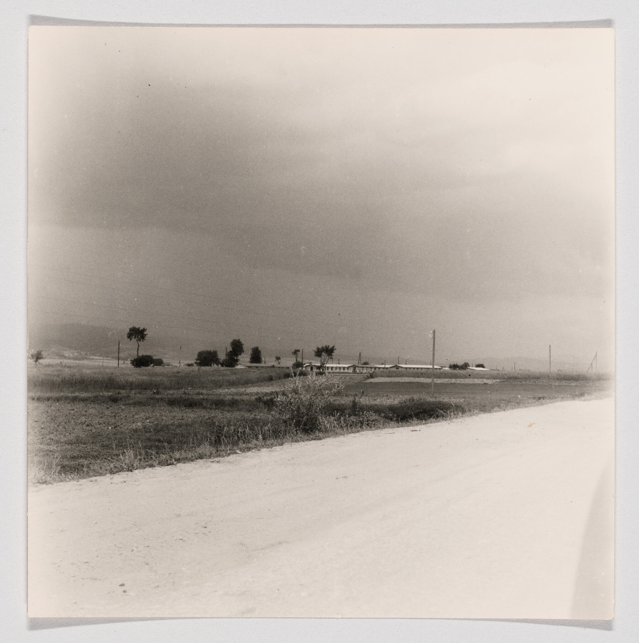 Dirt road runs past open fields and scattered trees toward low buildings under cloudy sky.
