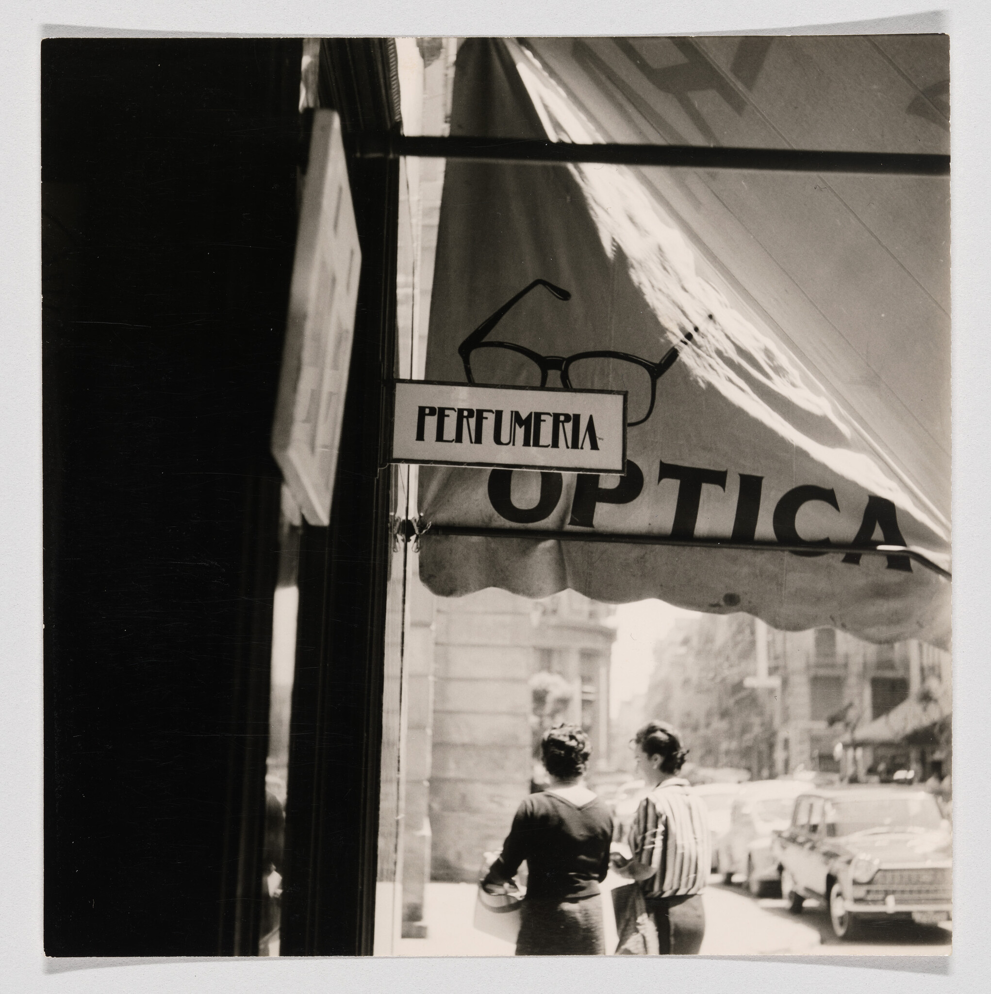 A street scene showing a perfumería and óptica awning above two women walking past vintage cars.