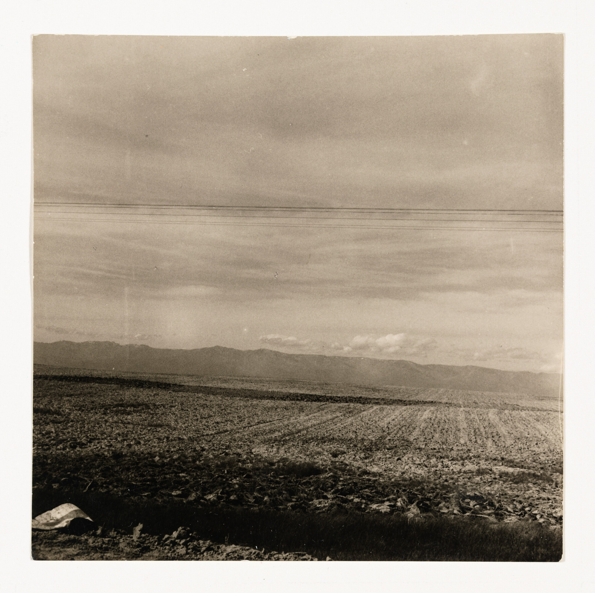 A wide plowed field stretches toward distant mountains under a cloudy sky with power lines.