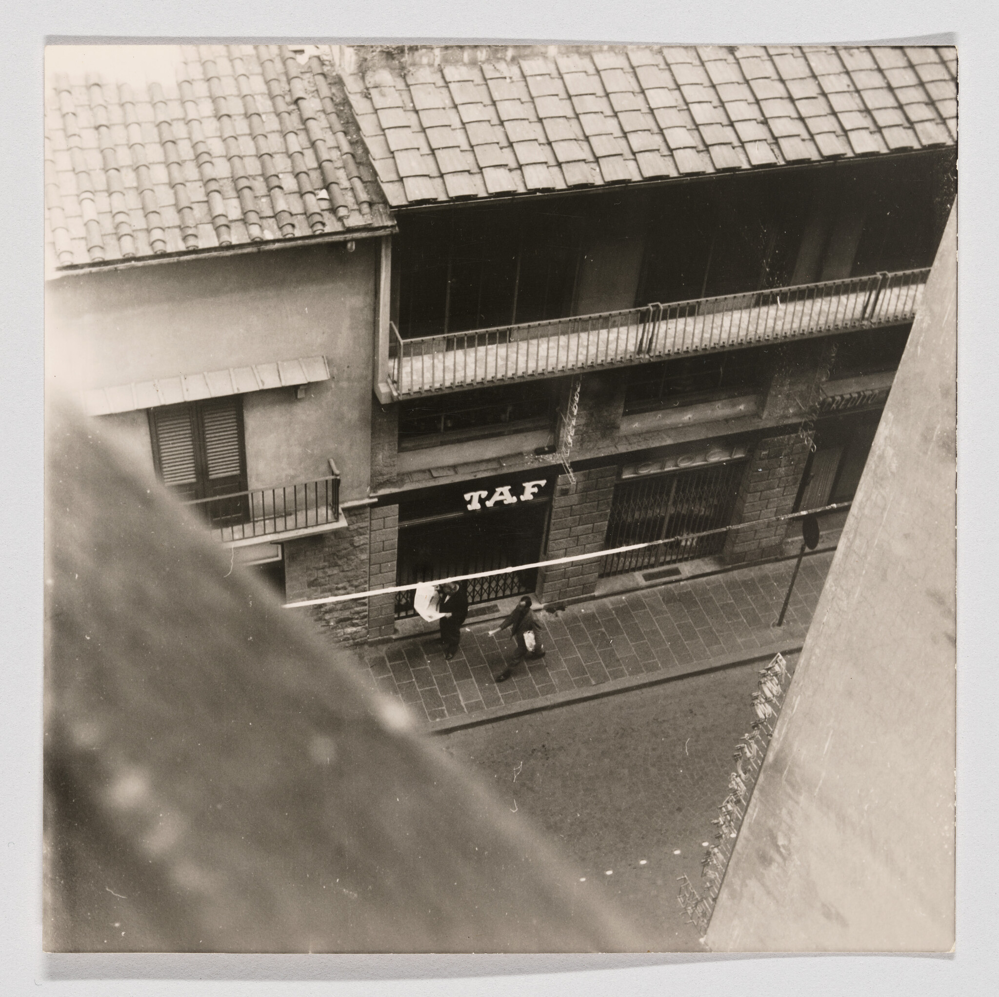 A black and white photograph taken from an elevated angle showing a street scene with two buildings facing each other. The building on the left has a tiled roof and shutters on the windows, while the building on the right features a balcony running along its length. Below the balcony, there is a sign with the letters "TAF" on the facade. Two people are walking on the sidewalk, and there is a streetlight and a utility pole visible. The edge of the roof in the foreground creates a diagonal line across the top left corner of the image.