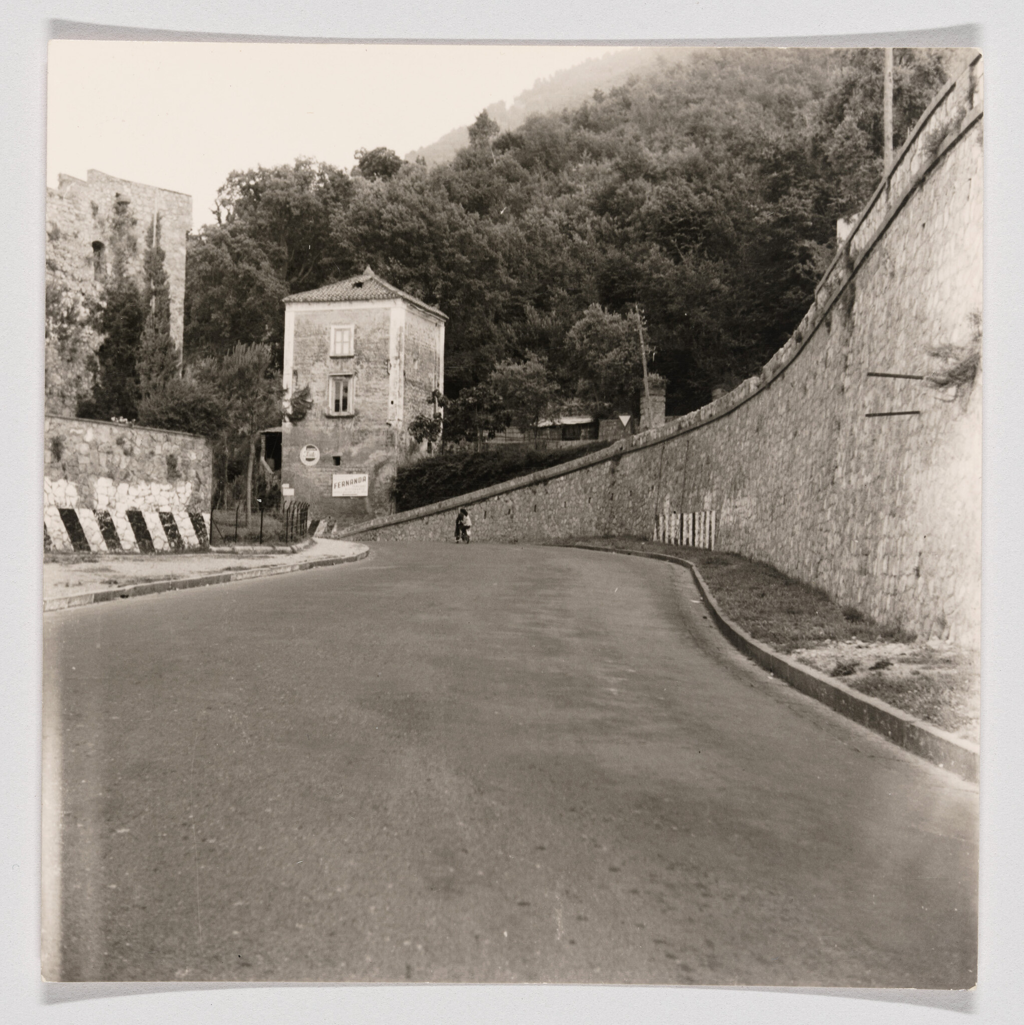 A lone person walks along a curved road beside a tall stone wall and old building.