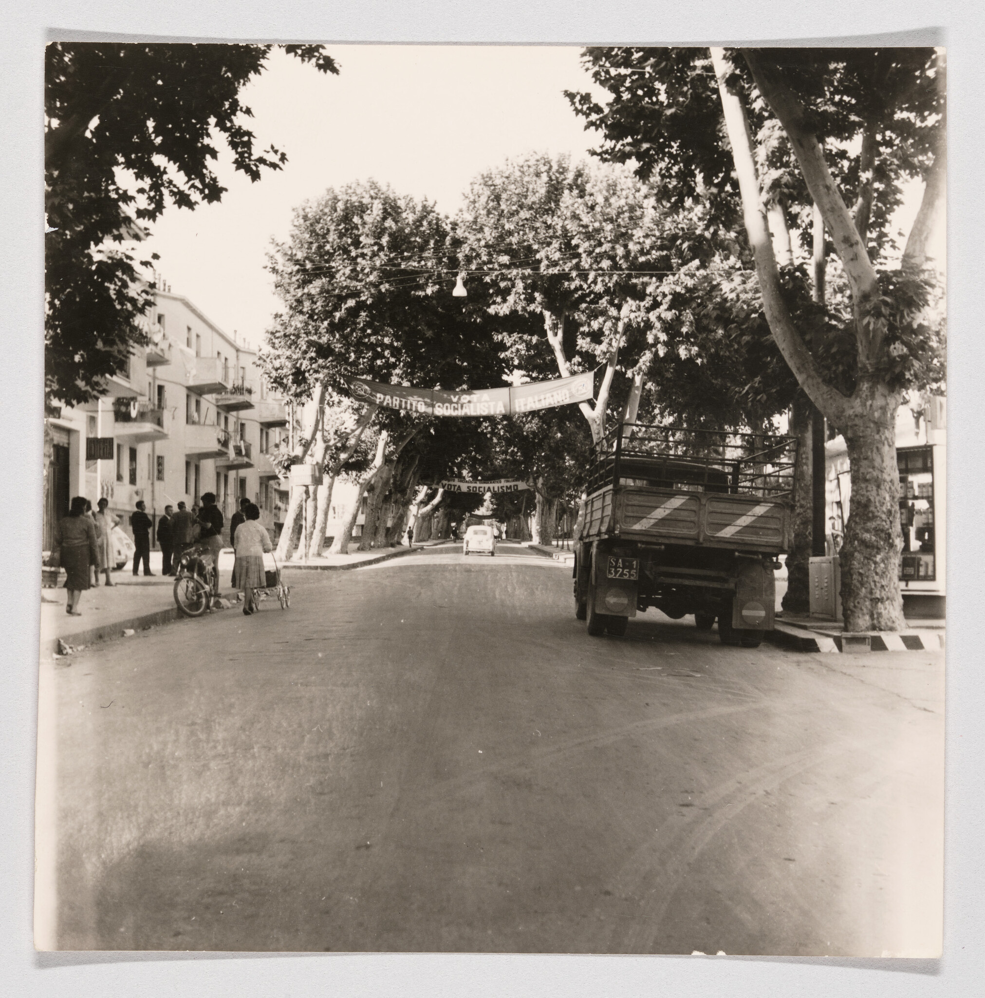 A tree-lined street with a parked truck, people on the sidewalk, and "Partito Socialista Italiano" banners overhead.