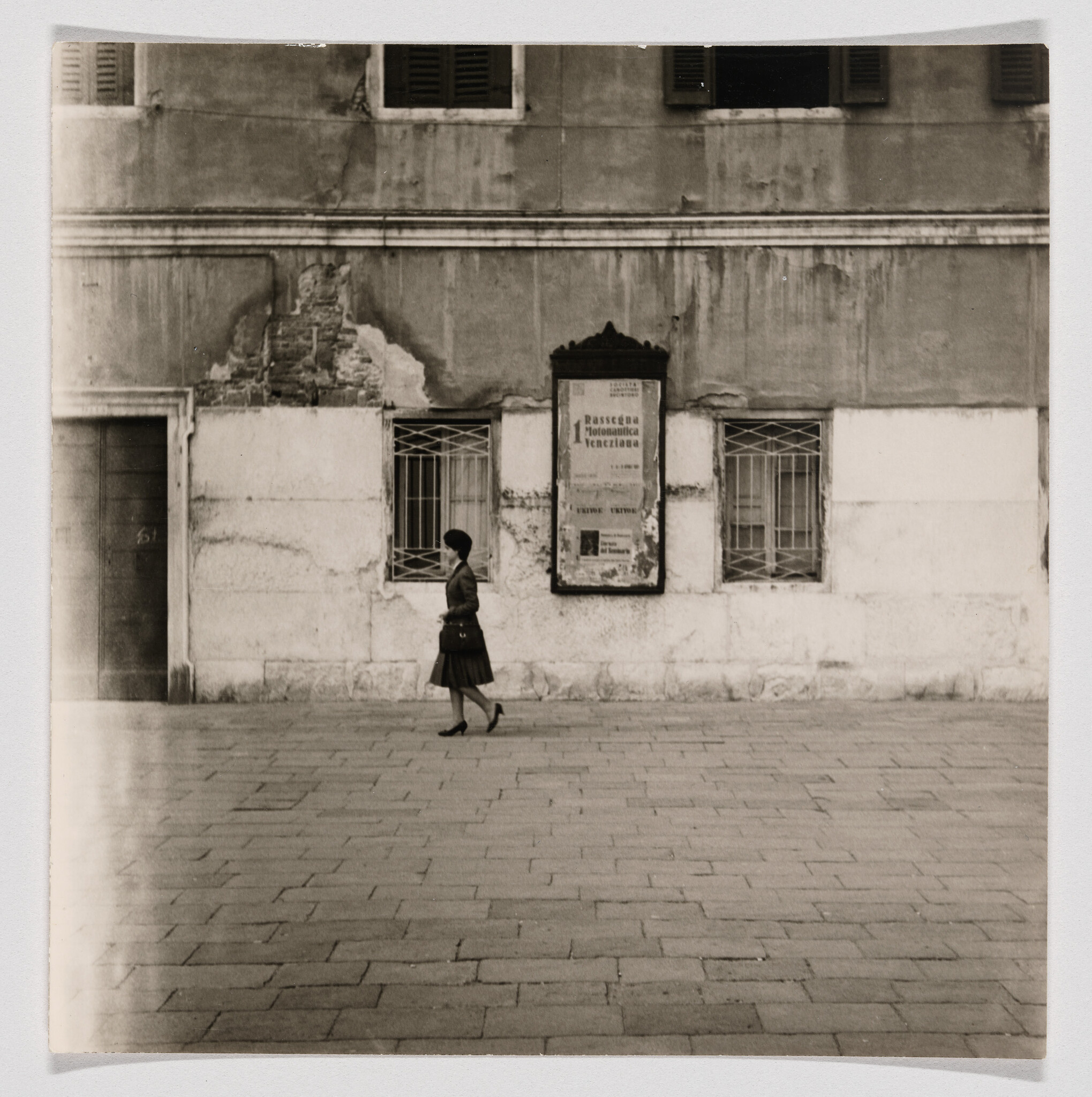 A woman in a skirt and heels walks along a paved plaza past a weathered building.