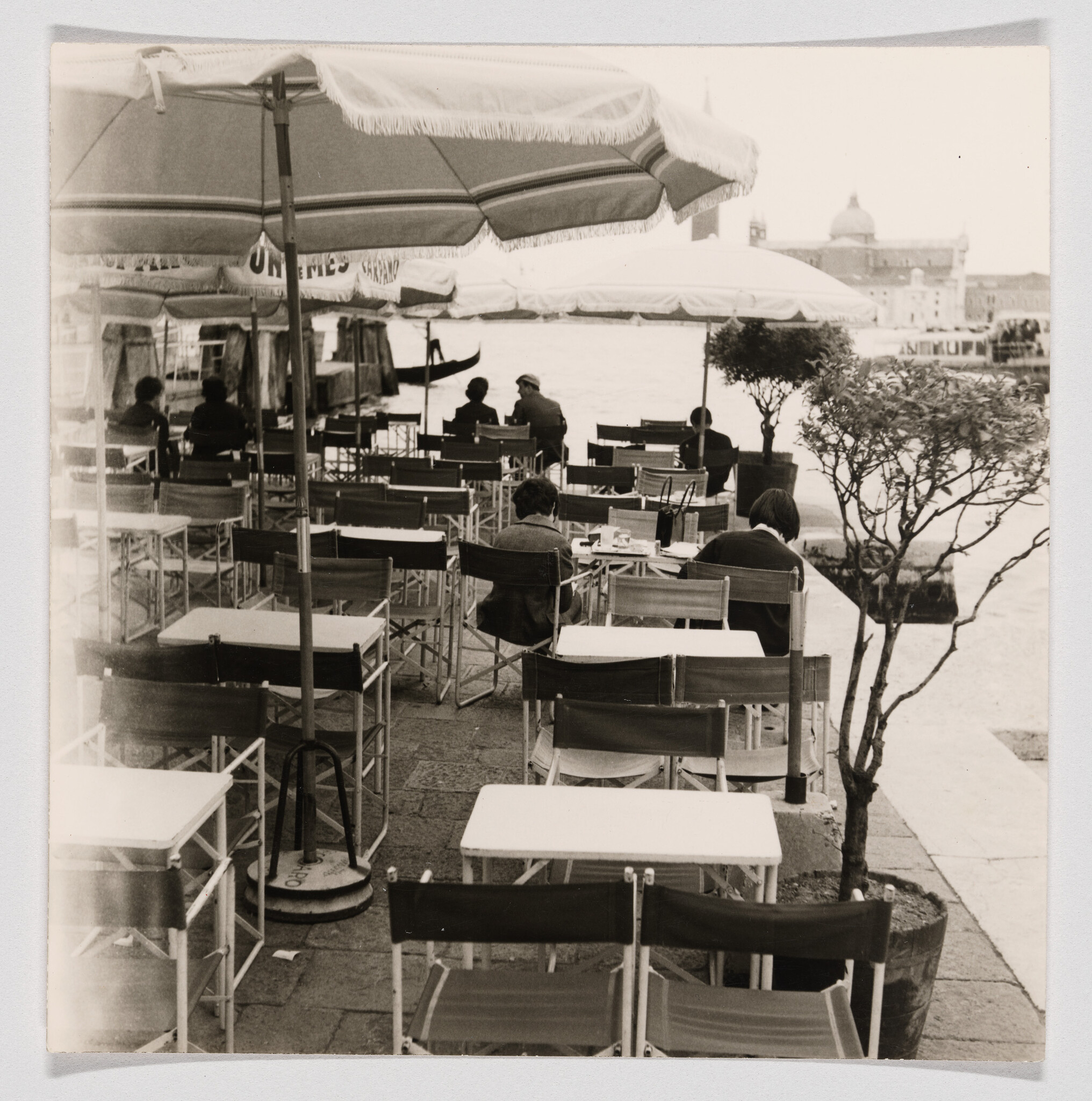 Empty café tables and chairs under umbrellas along a waterfront with a few seated patrons.