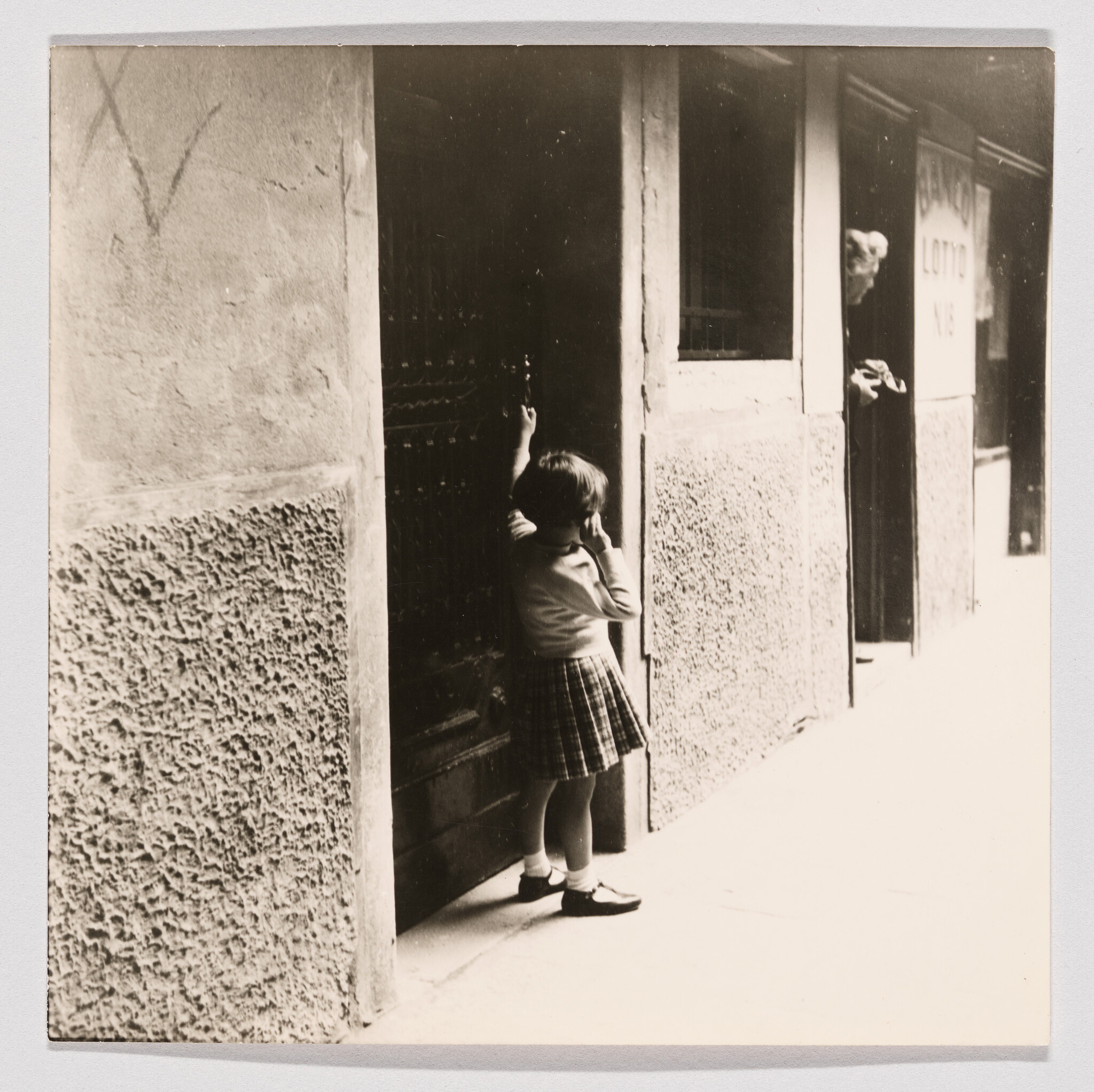 A young girl in a plaid skirt stands at a doorway holding the door while an older man peeks out.
