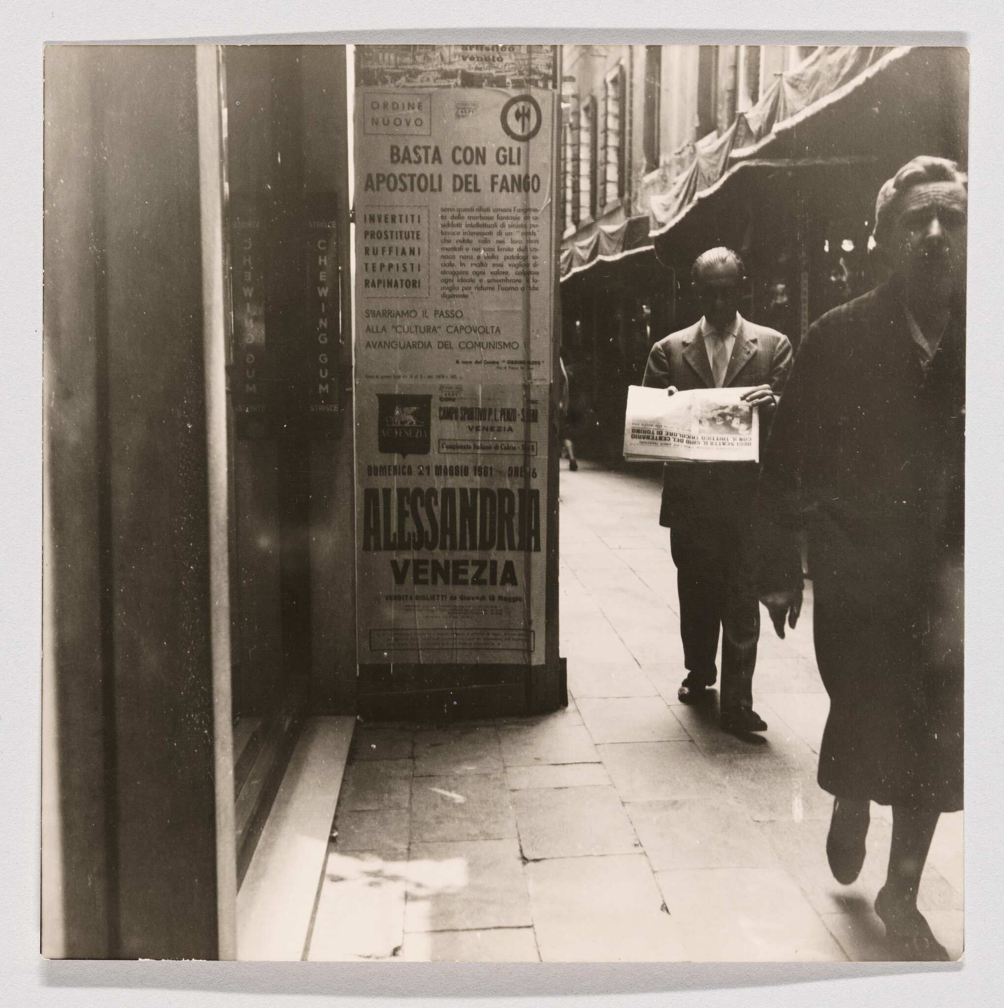 A man walks down a narrow Venetian arcade holding a folded newspaper near posters.
