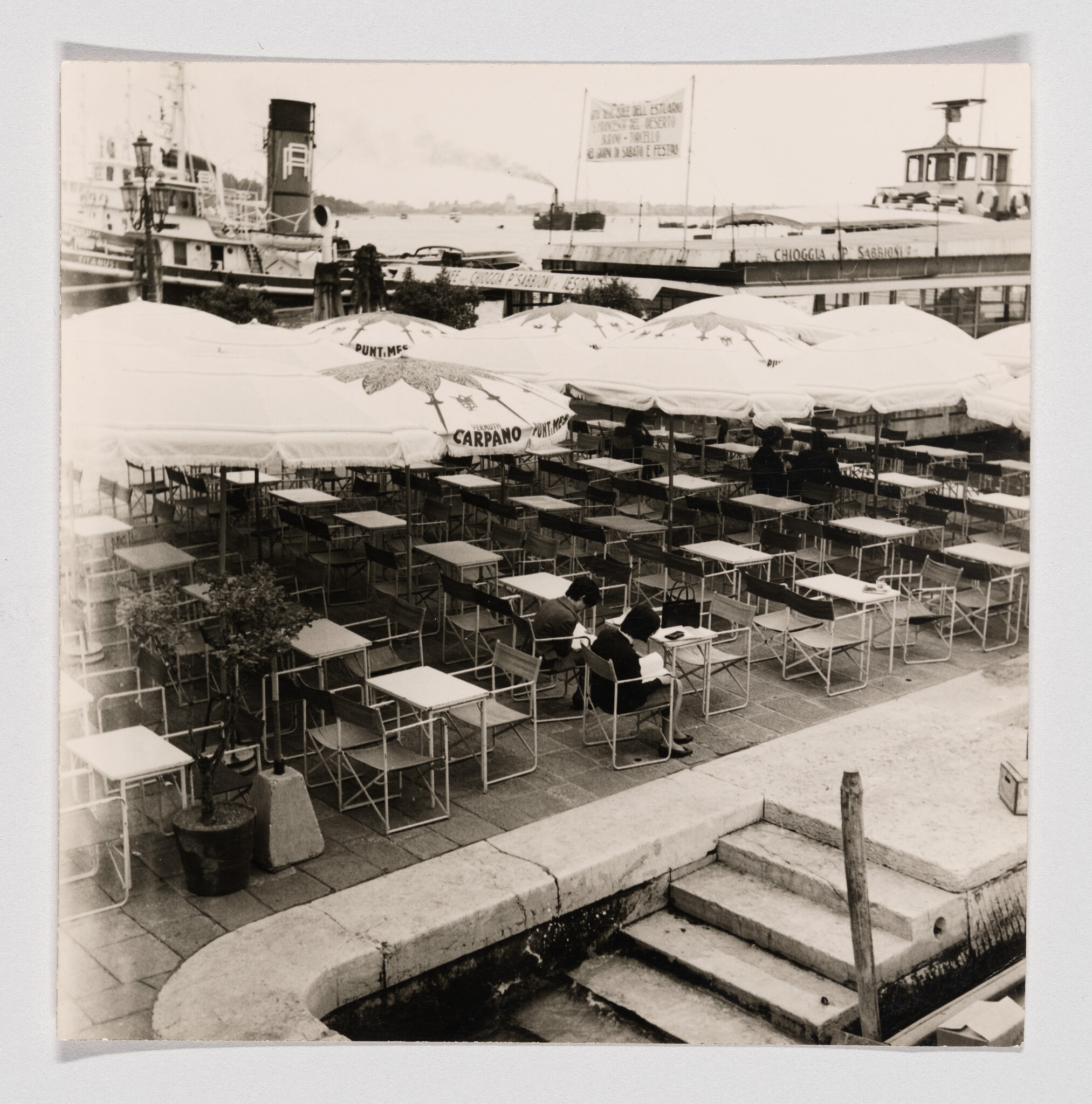 Empty café tables and umbrellas line a waterfront terrace while two people sit and read.